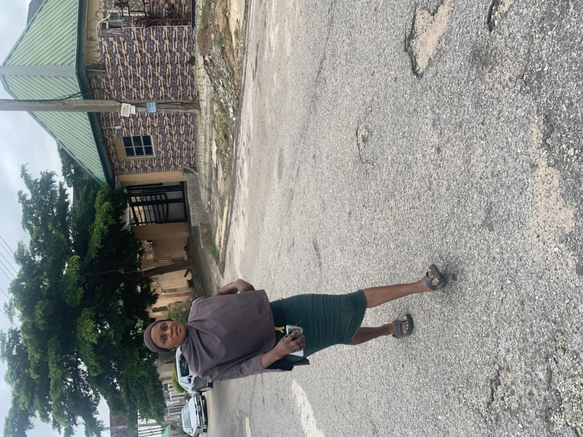 A woman is walking down a street in Abuja, Nigeria. She is wearing a dark grey wrap over a dark green skirt and dark grey sandals. She is holding a smartphone in her right hand. The street is paved with asphalt and shows signs of wear and tear, with several potholes visible. In the background, there are buildings, including one with a green metal roof and a brick-patterned facade. A large tree with green foliage is also visible. Cars are parked and driving on the street. The sky is overcast.