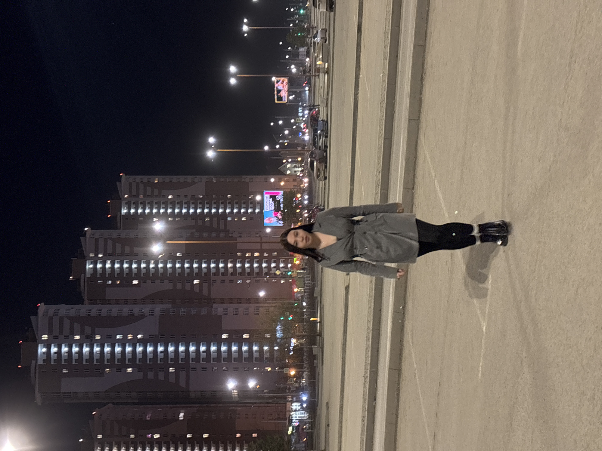 A sideways, full-length shot shows a woman standing on a pedestrian crosswalk at night in the city of Perm, Russia. She is dressed in black leggings and a gray belted coat. Tall apartment buildings with many lit windows can be seen in the background. Street lights and billboards illuminate the scene. The sky is dark. Cars are on the road in the background.
