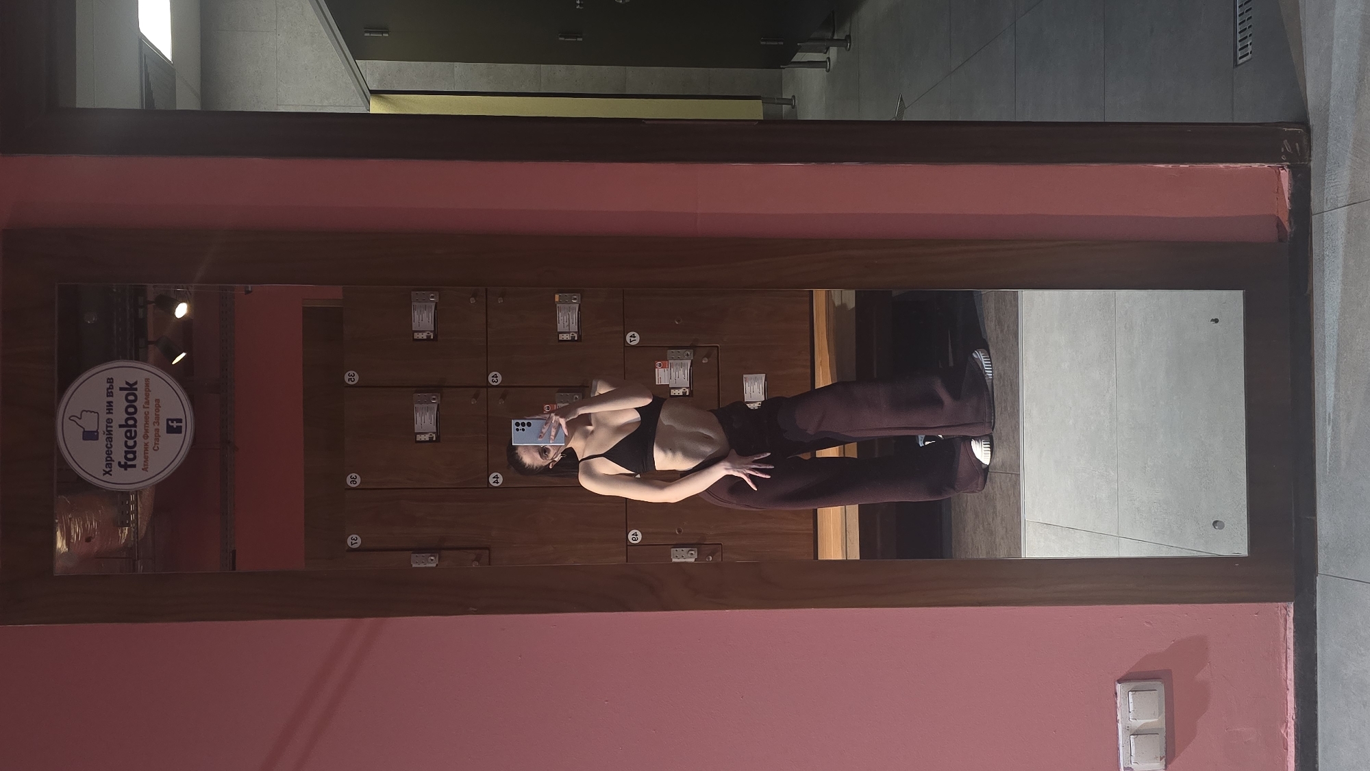 The image shows a young woman taking a selfie in a mirror. The mirror is framed with a pink border and reflects a wall with lockers. On the left side of the mirror is a Facebook logo, with the phrase "Харесайте ни във facebook," followed by "Атлетик Фитнес Галерия Стара Загора." The lockers are labeled with numbers, and the woman is wearing a black top and maroon pants. She is holding a blue smartphone to take the picture. The mirror reflects part of a gray tiled floor and wall. The location is the Athletic Fitness Gallery in Stara Zagora, Bulgaria.