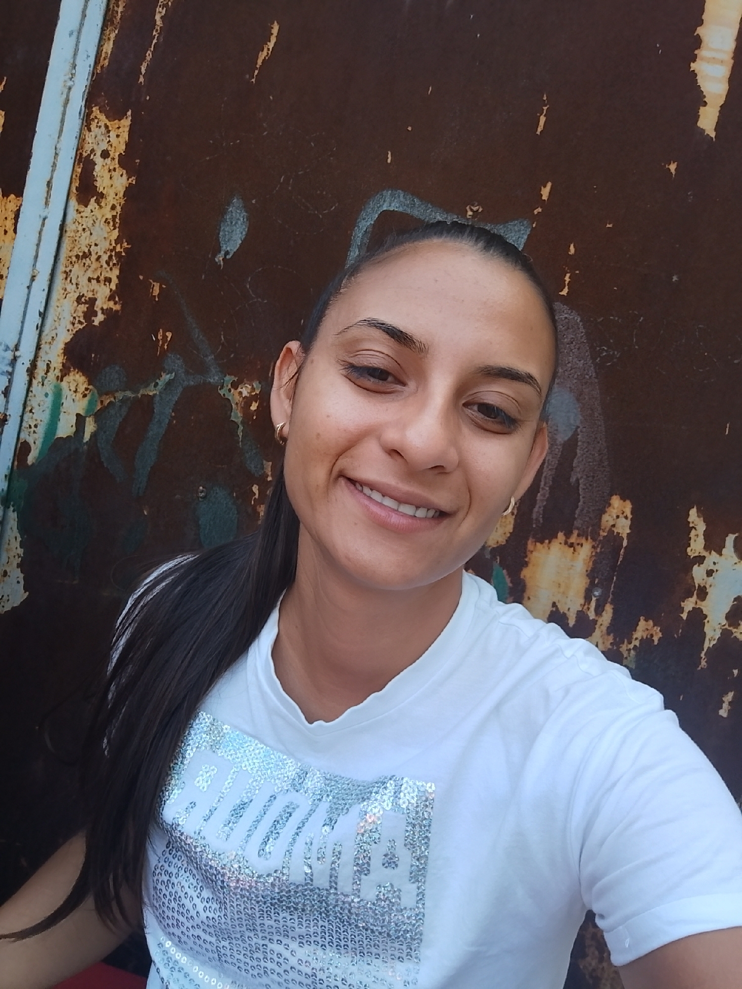 A young woman with dark hair pulled back in a ponytail, wearing a white t-shirt with silver sequins, smiles at the camera. She is positioned in front of a textured, rusty metal background with blue and white painted elements. The overall impression is a selfie taken outdoors. The location is specified as the city of Yambol, Bulgaria.