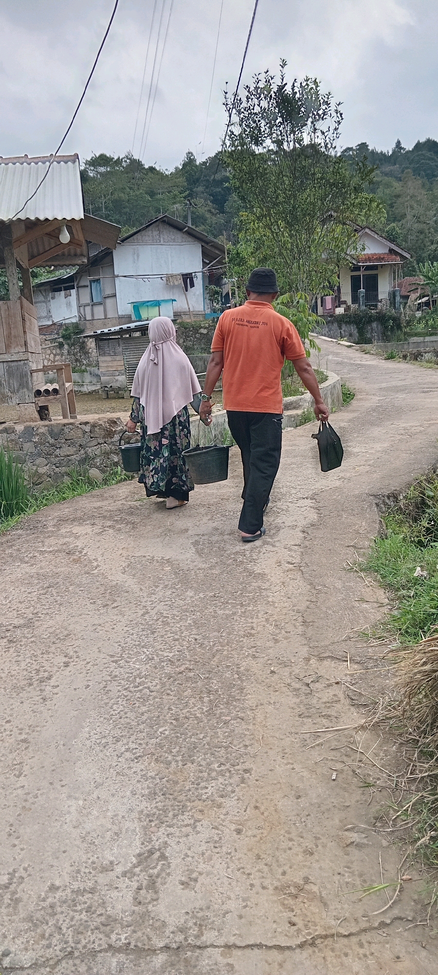 An overhead shot shows two people walking away from the camera on a paved road in Sodonghilir, Indonesia. The person on the left is a woman wearing a floral dress and a pale pink hijab, carrying two buckets, one in each hand. The person on the right is a man wearing an orange t-shirt and black pants, also carrying a black bag in his right hand. The road is surrounded by greenery and some houses, indicating a rural or semi-rural setting. The sky is overcast.