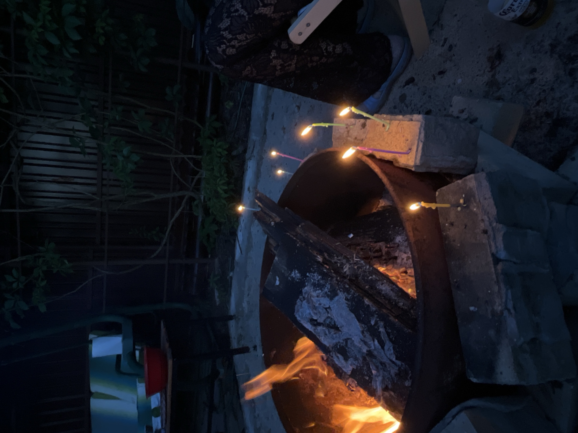 The image depicts an outdoor scene at dusk, possibly in a backyard or garden area, illuminated by a bonfire and several lit candles. The bonfire is contained within a large, dark metal fire pit and contains burning logs. Several thin, colored candles are stuck into bricks or small concrete blocks arranged around the fire pit, some appearing to be drippy with wax. In the background, a dark fence with vertical slats is visible, partially obscured by leafy branches. To the left, a portion of a table with a red bowl and what appears to be a chair or bench with blue and white cushions can be seen. On the right side of the image, the lower half of a person's legs is visible, wearing dark patterned trousers and light blue shoes. A portion of a white object, possibly a chair or table leg, is also present. The ground appears to be a mix of gravel and dirt. A bottle with a Corona Extra label is visible in the lower right corner. The overall atmosphere suggests a casual gathering or celebration, possibly a birthday, given the presence of candles. There is no specific indicator that this scene is located in Вельяминово, Russia.