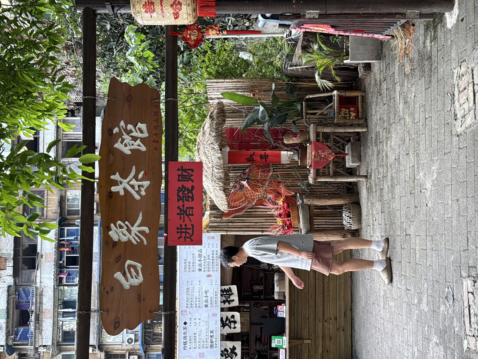 A medium shot depicts a street scene in the Yuzhong District of Chongqing, China.
A man stands on the right side, facing the storefront. He is wearing a light grey t-shirt, pink shorts, white socks, and beige slides. He appears to be looking at a menu posted outside a tea shop.
The storefront is decorated in a traditional Chinese style, with wooden signs featuring Chinese characters. A sign on the left reads "白象茶馆" (Bai Xiang Tea House), and another on the right reads "进者发财" (Those who enter become rich). There's also a detailed menu listing various teas and snacks with prices. The tea shop has a bamboo-style facade and a decorative thatched roof.
The street is paved with grey bricks. In the background, there are buildings with windows, and laundry hangs between some of them. Green plants are visible throughout the scene, adding a touch of nature to the urban setting.