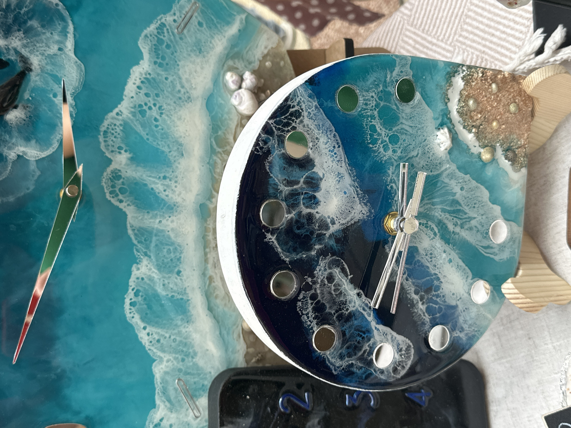 The scene depicts multiple artisan resin art pieces, primarily clocks, displayed in what appears to be an indoor commercial setting in the city of Unknown, Unknown. In the foreground, a round resin clock stands upright on a light wooden easel. Its face is patterned with a marine theme, featuring a gradient from dark blue to lighter turquoise, detailed with white swirling patterns representing ocean waves and foam. The bottom right segment of the clock face simulates a sandy beach with small, textured inclusions. Twelve circular depressions serve as hour markers; some are reflective silver, while others are dark green or black. Silver hour, minute, and second hands are present, indicating approximately 7:55. The clock has a white outer rim.
Behind this foreground clock, a larger, rectangular resin art piece, also depicting an ocean scene with turquoise water and white waves, is visible. This background piece incorporates clock mechanisms, with two stylized clock hands shaped like birds in flight, one tipped in red and the other in green, pointing towards roughly 10:45. Small white spiral shells are scattered on the sandy portion of this larger artwork. To the left of the foreground clock, a segment of a black object featuring embossed blue numbers "2" and "3" is partially visible. The foreground clock rests on a light-colored, textured fabric surface. A brown and white patterned textile is partially visible on the far right. No individuals are present within the frame.