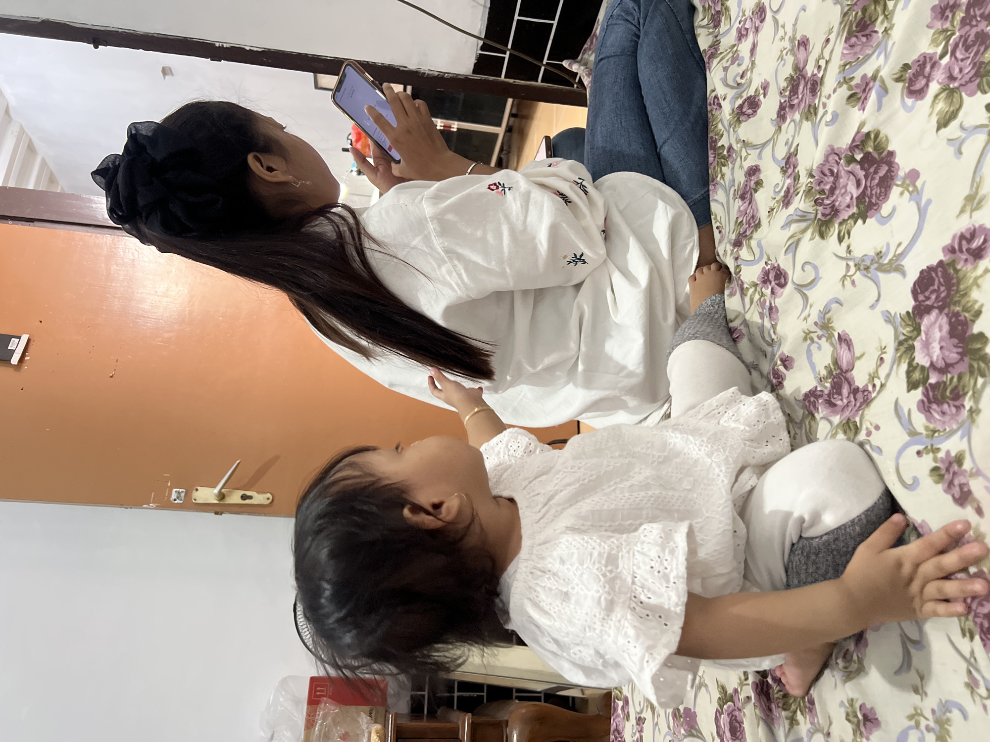 In Yogyakarta, Indonesia, on a bed covered with a floral-patterned comforter, a young child and a teenager are seen in a domestic setting. The child, facing away from the camera, is wearing a white, lace-trimmed shirt and white pants with gray leggings. The teenager, also facing away, has long dark hair tied up with a black scrunchie and is wearing a white shirt with embroidered flowers and blue jeans. She is holding a smartphone and appears to be looking at it. The background shows a closed wooden door and a white wall.