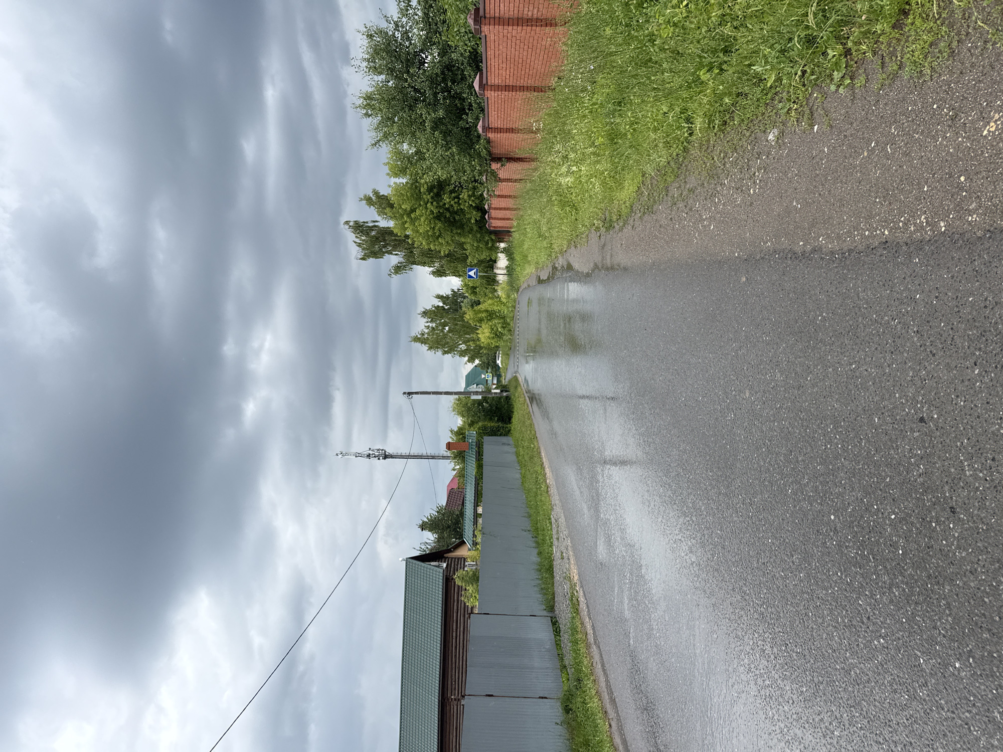 The image depicts a rural road in Kazan, Russia, on a cloudy day, likely after rainfall as the asphalt is wet and reflective. The road curves to the left, leading into the distance. On the left side of the road, a grey corrugated metal fence runs along the edge, behind which are wooden houses with green roofs. A power line stretches across the sky, supported by a utility pole. A tall communication tower is visible further down the road. On the right side, a red brick fence borders a grassy area with lush green vegetation and trees. A blue and white road sign, indicating a pedestrian crossing, is also present on the right. The sky is overcast with dark grey clouds. There are no people or vehicles visible in the image.
