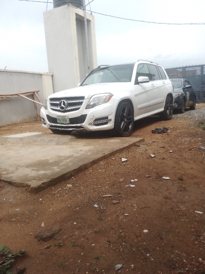 A white Mercedes-Benz ML-Class SUV is parked on a concrete driveway. The vehicle is facing the camera, parked on the right side of the driveway. The front wheels are turned slightly to the right, and the car is parked on a slight incline. The car has black wheels and tires, and the tires are partially obscured by the car's body. There is a metal gate on the right side of the car, partially obscured by the SUV. There is a building in the background on the left side of the car, and some unpaved ground on the right. The ground is covered in dirt, pebbles, and some debris. The location is in Bida, Nigeria, at the coordinates latitude 9.045, longitude 6.003.