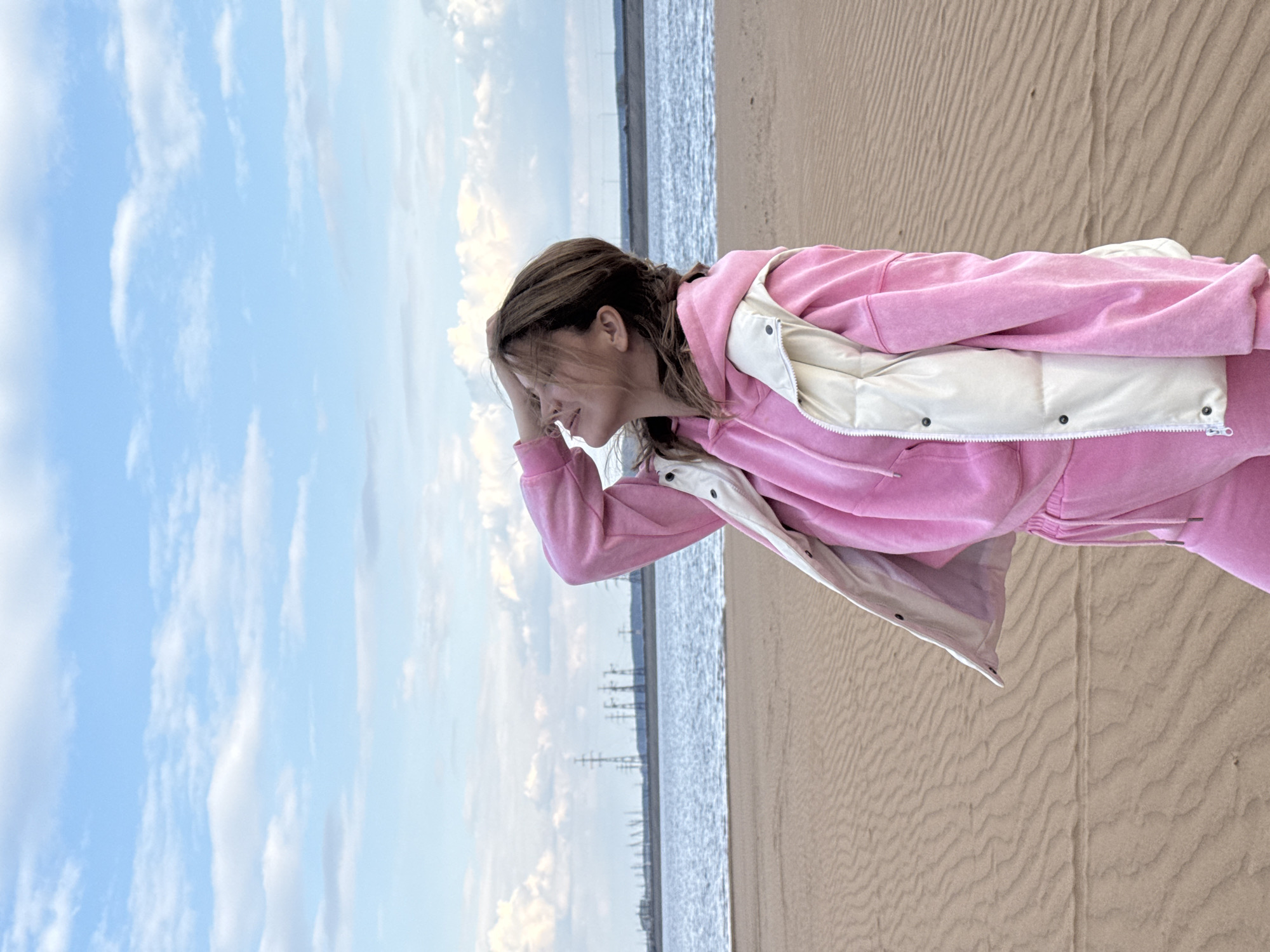 A woman stands on a sandy beach, shielding her eyes with her hand, possibly looking out at the water or horizon. She is wearing a pink velour tracksuit with a white puffer vest. Behind her are power lines running along the water, suggesting an industrial or urban area near the beach. The sky is blue with scattered white clouds. The location is Noviy, Russia, based on the visible power lines and beach environment. The ground appears to be sandy with textures indicative of windblown sand.
