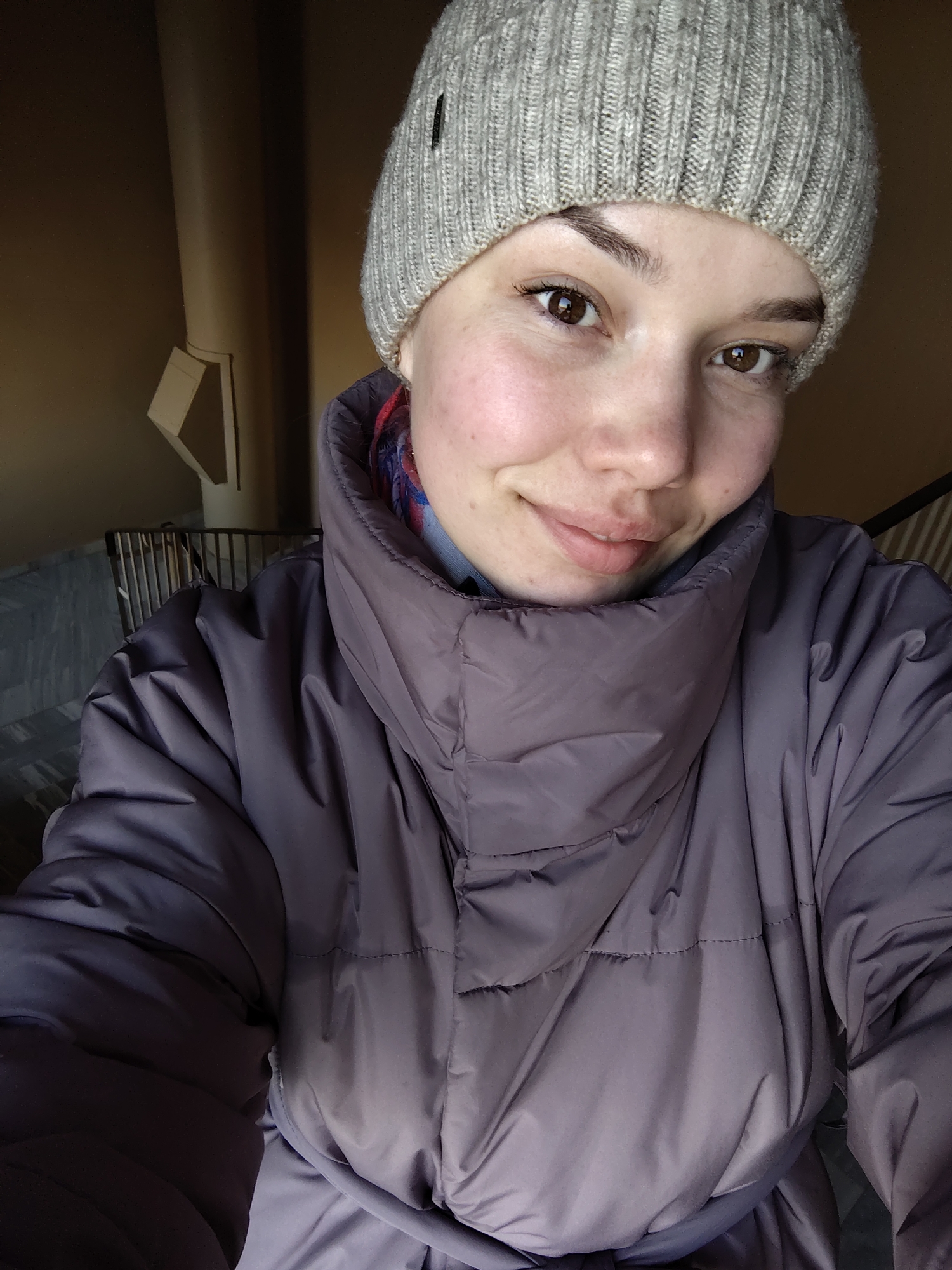 A close-up, frontal photograph depicts a young adult female situated within an indoor environment in the city of Марковский, Russia. The subject is wearing a light gray knitted beanie and a dark purple puffer jacket with a prominent high, standing collar. A segment of a multicolored garment, displaying red and blue patterns, is visible beneath the jacket's collar. She possesses light skin, brown eyes, and dark eyebrows, and exhibits a subtle smile directed towards the camera. The background is out of focus but reveals a warm brown wall, a light-colored vertical pipe or conduit with a triangular bracket to the left, and a dark metal railing partially obscuring a patterned tiled floor, suggesting a stairwell or common area within a building.