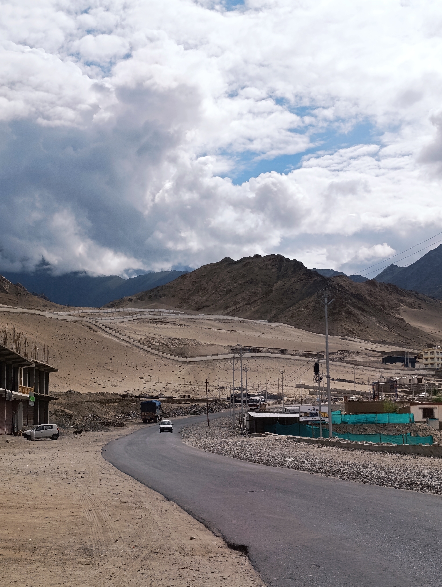 A landscape image captures a road winding through a barren, hilly terrain under a partly cloudy sky. The scene appears to be in or near the city of Leh, India, known for its mountainous landscape and Buddhist monasteries.
On the left side of the road, a two-story building, possibly under construction, stands with exposed concrete and some scaffolding. A white car is parked in front of the building, and a dark-colored dog is visible near it. The road curves into the distance, with two white cars and a truck driving on it. The terrain is mostly dry and sandy, with sparse vegetation.
In the background, rolling hills and mountains dominate the landscape. A winding path, possibly a staircase or a road, with white railings, ascends one of the hills. The sky is filled with dramatic clouds, some dark and heavy, suggesting an approaching storm, while others are lighter and brighter. A few utility poles with wires are scattered along the roadside, indicating a developed area. Buildings, some appearing to be residences or small businesses, are visible in the distance to the right, suggesting the presence of a settlement. The overall impression is one of a remote, arid, and mountainous region.