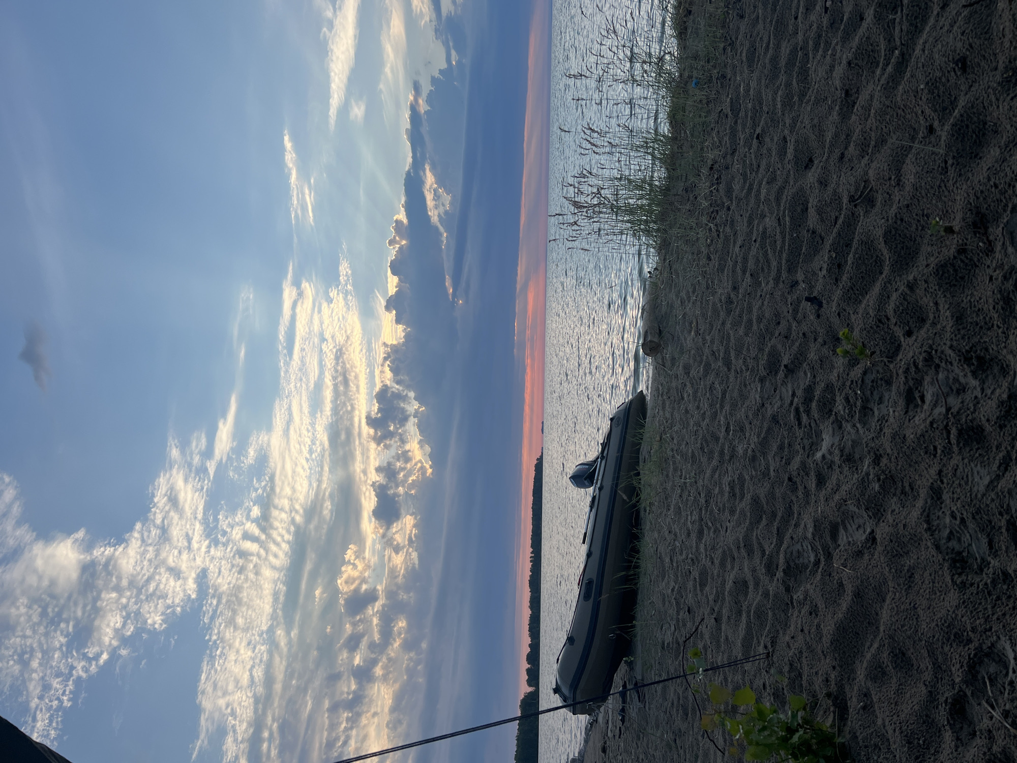 An inflatable boat is resting on the sandy shore of a body of water under a cloudy sky with visible sunbeams. The boat is dark green and appears to have a small outboard motor attached. Tall grasses grow along the water's edge. The sky transitions from light blue with white clouds to darker, dramatic clouds tinged with pink and orange near the horizon, suggesting either sunrise or sunset. The water is calm and reflects the sky. In the distance, a tree line is visible on the horizon, and a ship is present on the water far to the right. The sand in the foreground is textured with ripples and a few small plants. There are no people or animals visible in the scene. The location is the city of Borovoe, Russia.