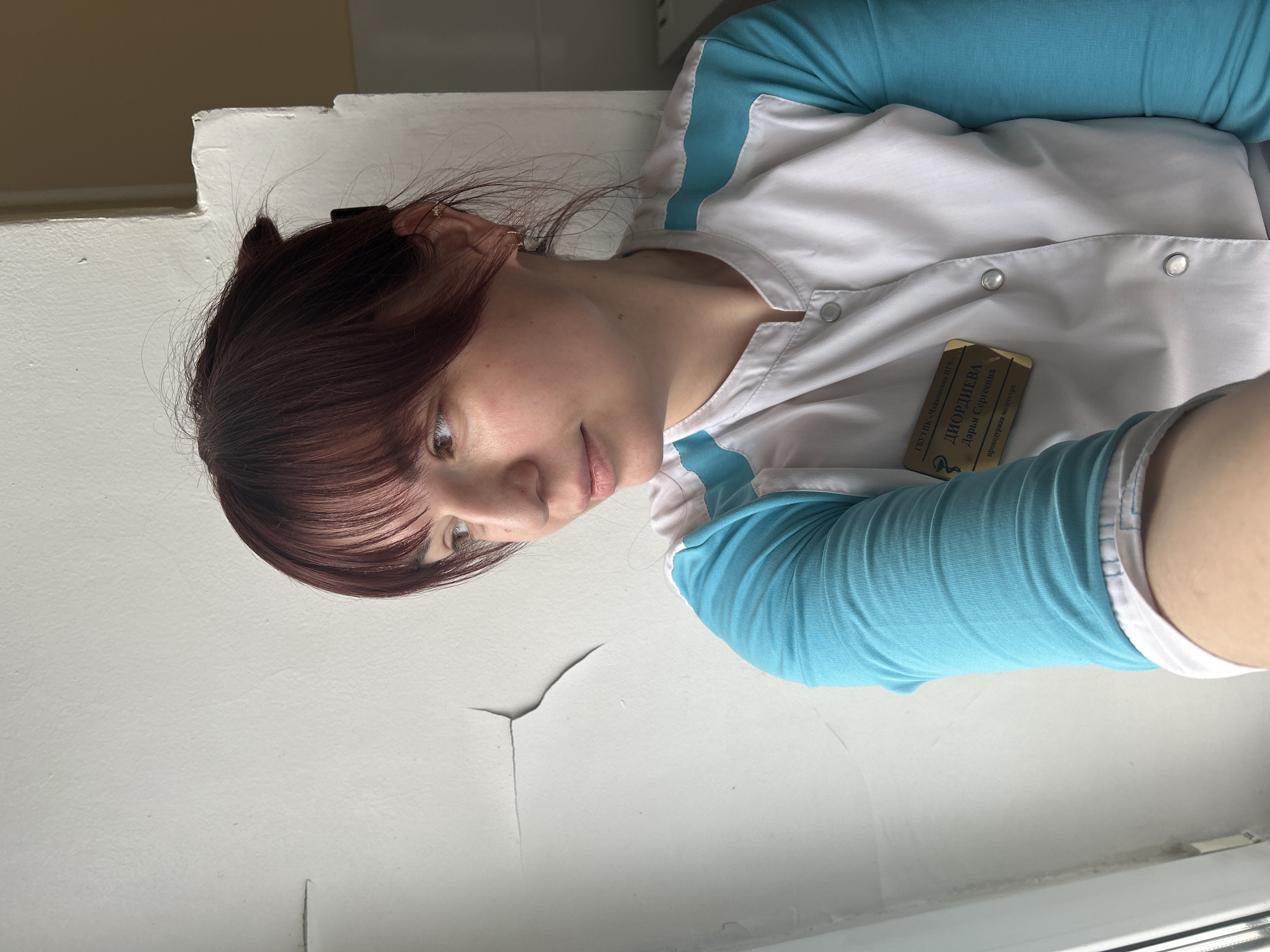 A woman wearing a medical uniform is depicted in a portrait-style image, taken indoors. She has dark brown hair with bangs and appears to be looking down towards the camera with a neutral, slightly contemplative expression. She is dressed in a light blue long-sleeved shirt or scrub top, covered by a white medical jacket fastened with snap buttons.
A gold-toned name tag is clipped to her right side. The text on the name tag, in Russian, identifies her:
- Top line: "ГКПБ им. Чанковской П.П." (likely a medical facility, possibly a State Clinical Psychiatric Hospital named after P.P. Chankovskaya)
- Name: "ДИОРДИЕВА Дарья Сергеевна" (Diordieva Daria Sergeevna)
- Role: "процедурная медсестра" (procedural nurse)
A small, medical-related logo is also visible on the badge.
The background consists of a plain, light-colored wall, showing visible signs of wear with peeling paint and cracks. The lighting is bright and even, suggesting a daytime setting. A small, delicate earring is visible in her left ear, and part of her hair is pulled back and secured with a dark clip. The image captures what appears to be a moment in a medical or administrative setting in Фоки, Russia.