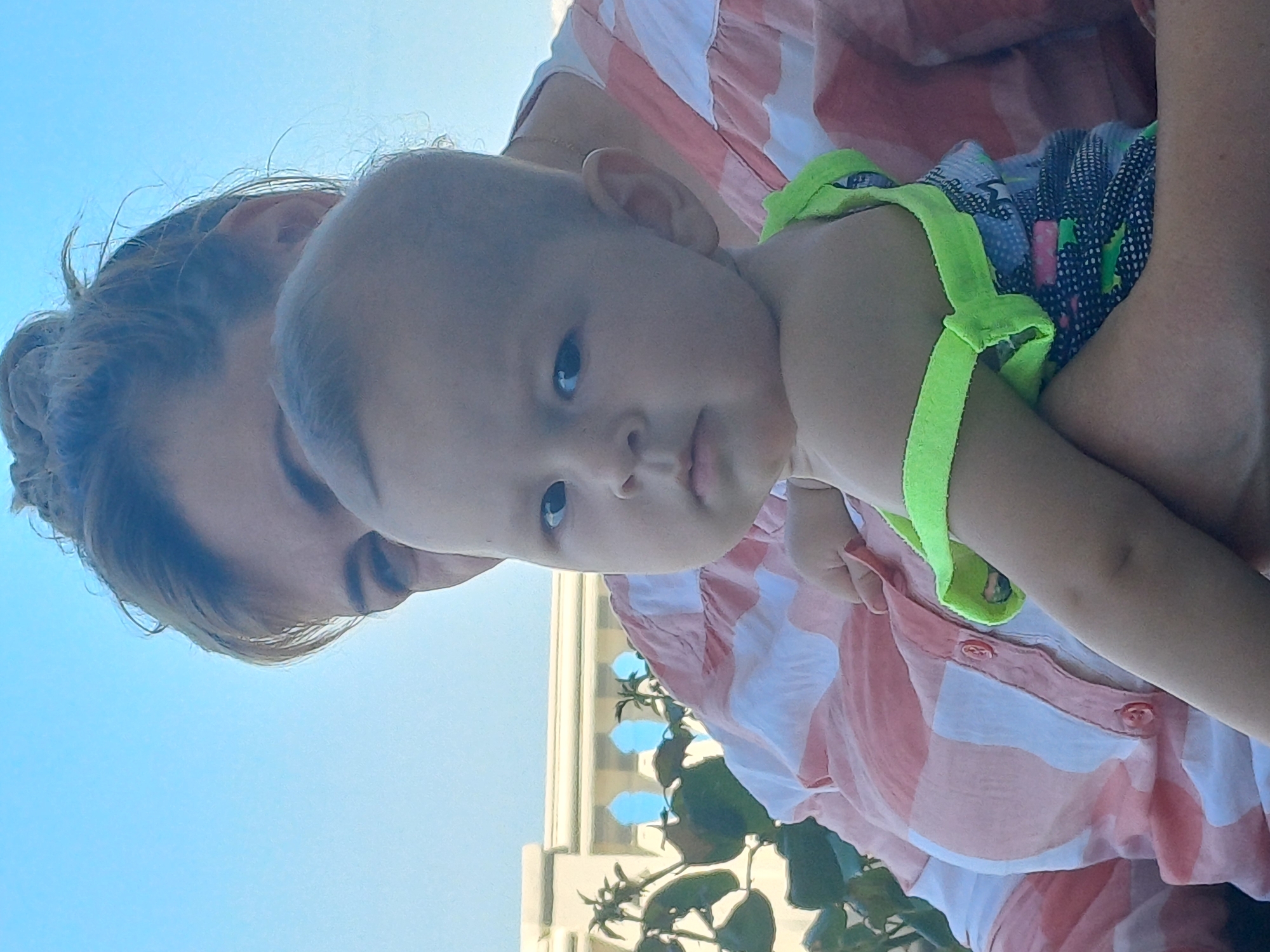 An outdoor, eye-level, medium shot captures a baby being held by an unseen adult. The baby, facing the camera with a serious expression, is positioned in the foreground. The baby's body is partially obscured by the adult's arms and chest. The adult is wearing a pink and white striped shirt. Behind them, a light-colored building with arched openings and lush green foliage can be seen under a clear blue sky. The architectural style of the building suggests it may be located in Sochi, Russia, possibly a resort or a decorative facade. The image has a bright, natural light, indicative of daytime.
