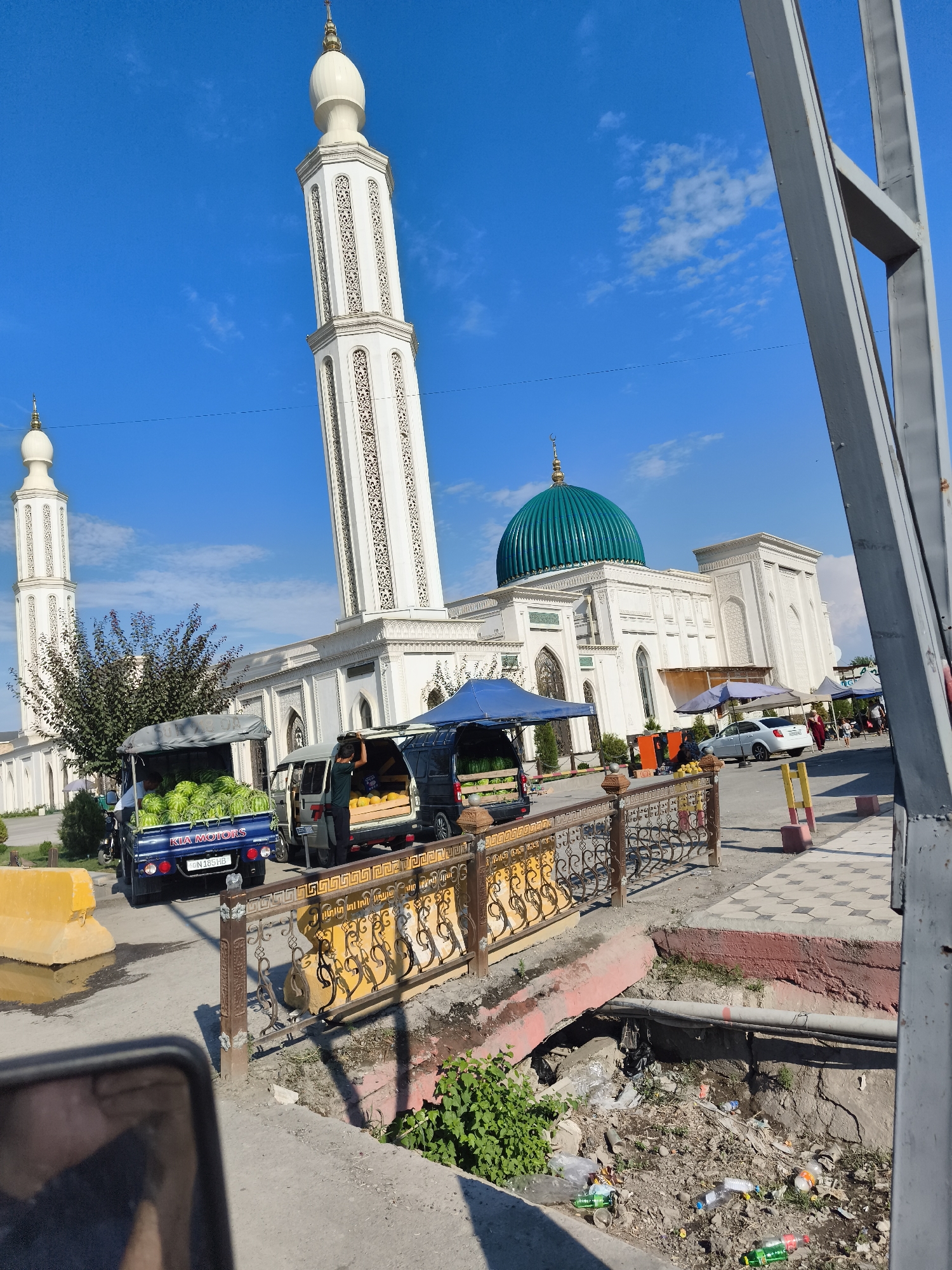 The image depicts an outdoor scene in Buz, Uzbekistan, likely during daytime given the bright sunlight and clear blue sky with scattered clouds. The primary subject is a large, white mosque with ornate minarets and a prominent green dome, characteristic of Islamic architecture.
In the foreground, a street is lined with vendors selling produce from the back of parked vehicles. One blue Kia Motors pickup truck is filled with watermelons, while a smaller van next to it displays what appear to be melons or similar fruits. A person is seen attending to the fruit in the van. Tents or awnings are set up over some of the vendor stalls, providing shade.
A decorative metal fence with intricate patterns separates the street from a lower area that contains a considerable amount of litter, including plastic bottles and bags, indicating a potential problem with waste management in the vicinity. A yellow concrete barrier is also visible on the left side of the road. A white car is parked near the mosque, and other people are seen in the background, suggesting a public area with ongoing activity. The overall atmosphere appears to be that of a bustling market day in a significant religious and public space.