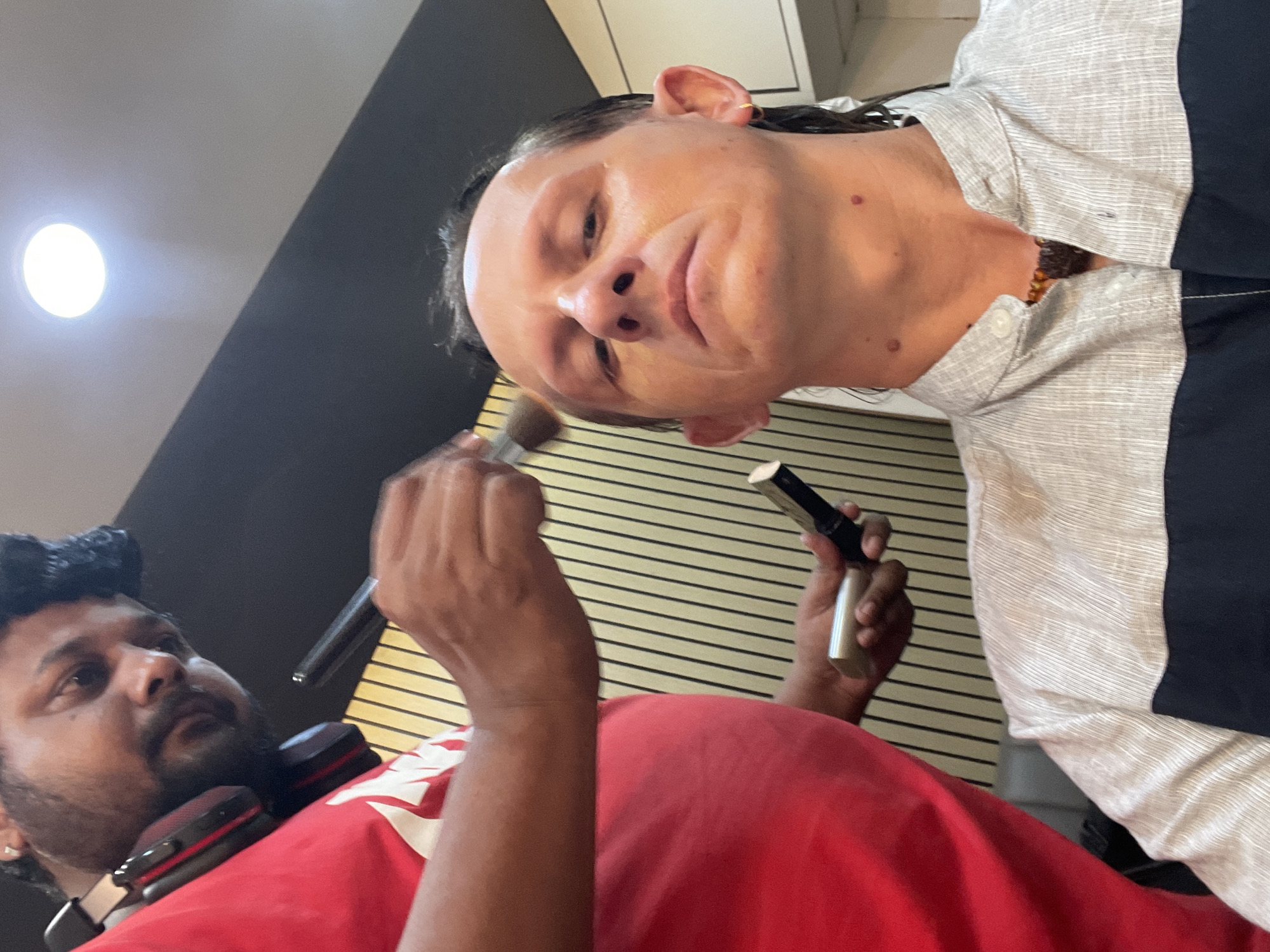 A man in a red t-shirt with headphones around his neck is applying makeup to another man's face with a brush. The man receiving makeup is wearing a light-colored shirt with dark trim and a beaded necklace. The background appears to be a recording studio with soundproofing panels. This scene is taking place in Mumbai, India.