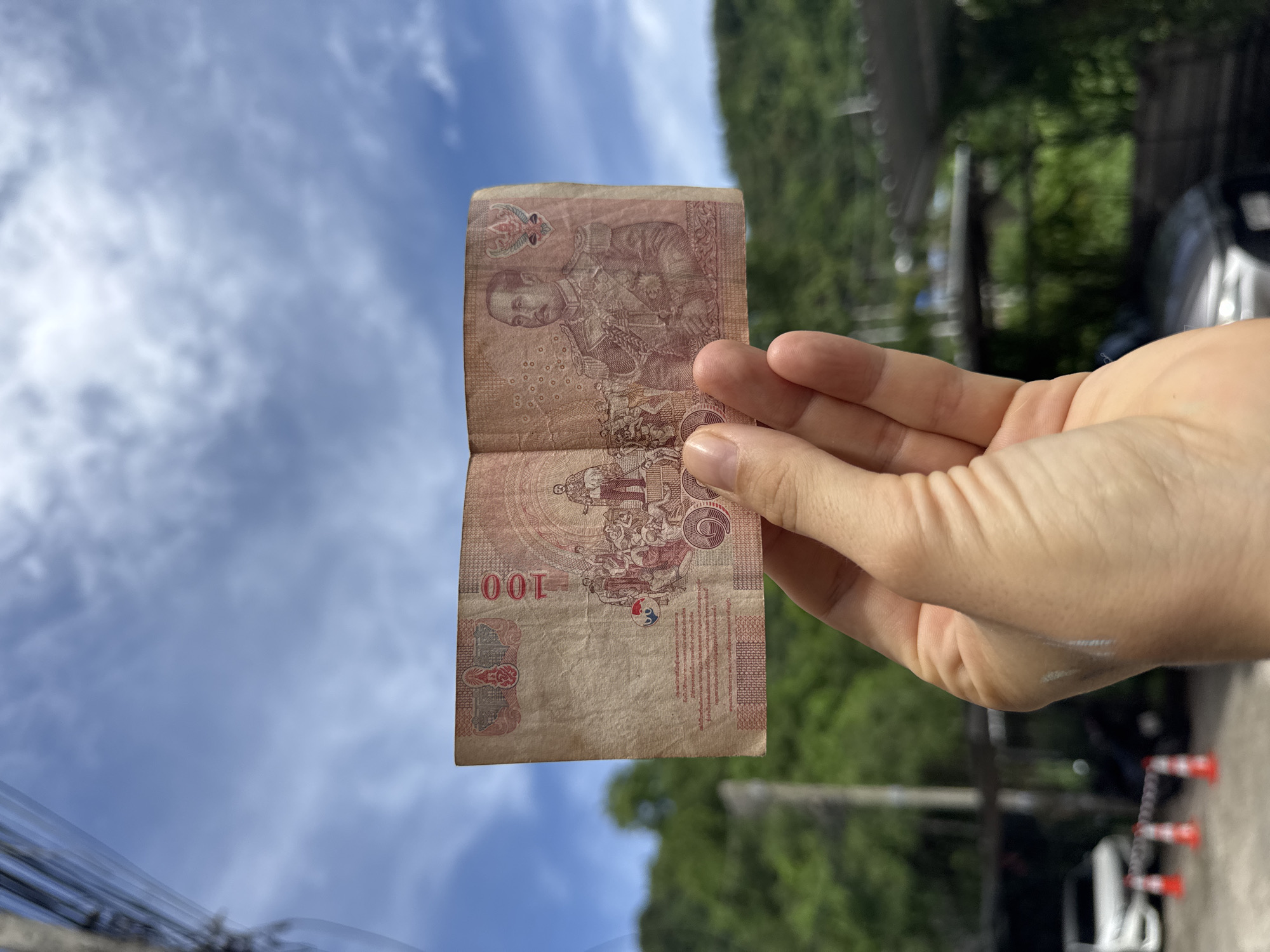 A person's hand holds a partially unfolded Thai 100 Baht banknote against a bright blue sky with scattered clouds. The banknote, printed in reddish-brown ink, features a portrait of King Bhumibol Adulyadej on the right side. The numeral "100" is prominently displayed on the left. Text in Thai script is visible on the lower portion of the banknote. In the background, out of focus, are lush green trees covering a hillside, suggesting a tropical environment. Below the trees, a glimpse of a paved area with parked vehicles and orange traffic cones can be seen, possibly indicating a street or parking lot in Karon, Thailand. The overall lighting suggests it is daytime.