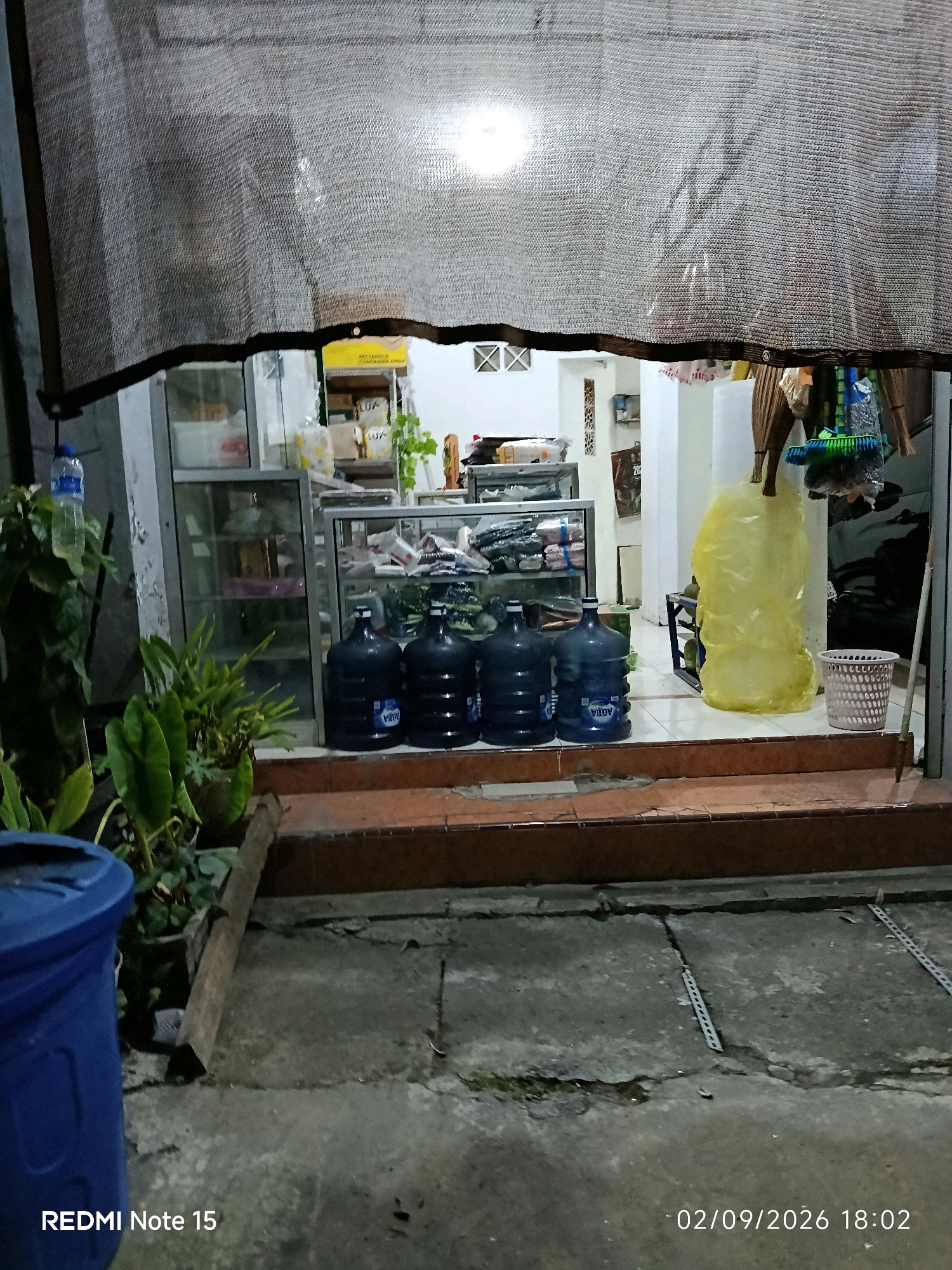 The image depicts the exterior of a small store or kiosk in Keboansikep, Indonesia. A large, coarse, greyish-brown fabric awning with visible grommets is pulled down, partially obscuring the view into the store. The awning appears to be made of a mesh material, allowing some light to filter through. Below the awning, a store window is visible, revealing shelves stocked with various items. In front of the store, several large blue water cooler jugs are arranged on the ground. To the left of the jugs, a green leafy plant in a pot is visible, and to the far left, a portion of a blue trash bin is in the foreground. To the right of the water jugs, a yellow plastic bag stands upright, and a white basket with a circular hole pattern is also present. The ground in front of the store is paved with concrete that shows some cracks and discoloration. The lighting suggests it is either dusk or nighttime, with artificial light sources visible inside the store, illuminating the awning from behind. The bottom of the image contains a watermark that reads "REDMI Note 15" and a date and time stamp "02/09/2026 18:02".