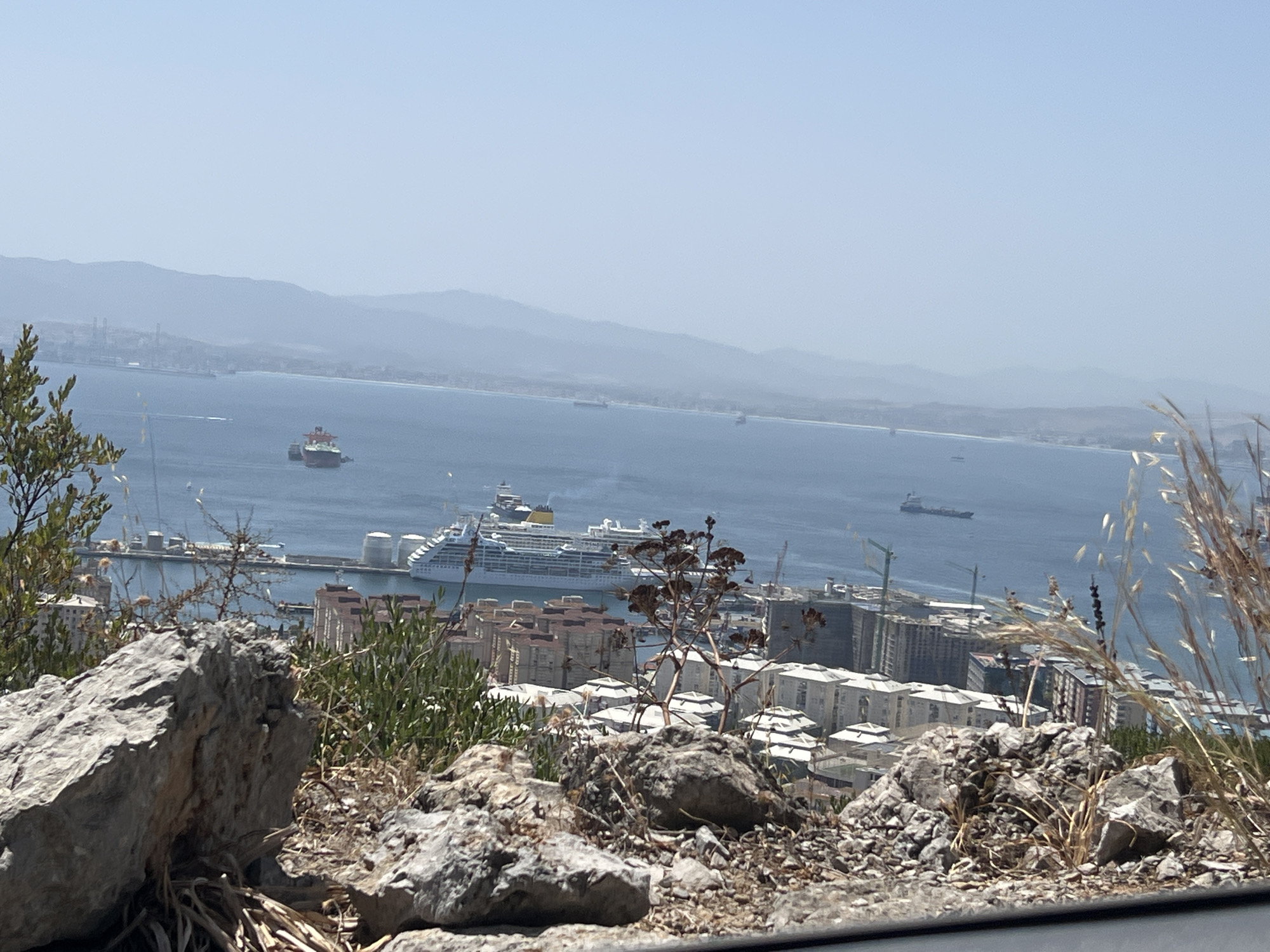 The image is a high-angle view of the city of Gibraltar, Gibraltar, taken on a clear, sunny day. In the foreground, large grey rocks and dry vegetation are visible, suggesting a natural hillside or elevated viewpoint. The midground shows a densely packed urban area with buildings of varying heights and architectural styles, some appearing residential and others commercial or industrial. Several buildings have light-colored facades and red or grey roofs.
Beyond the city, a large blue body of water, likely the Bay of Gibraltar, dominates the scene. Several vessels are present on the water. A large white cruise ship with multiple decks and yellow accents is prominent, docked or anchored near the shore. Other ships, including what appears to be a cargo ship and smaller boats, are also visible. In the distance, hazy, layered hills or mountains form the horizon line. The sky is a pale, clear blue. The overall impression is of a coastal city with significant maritime activity and a mountainous backdrop.