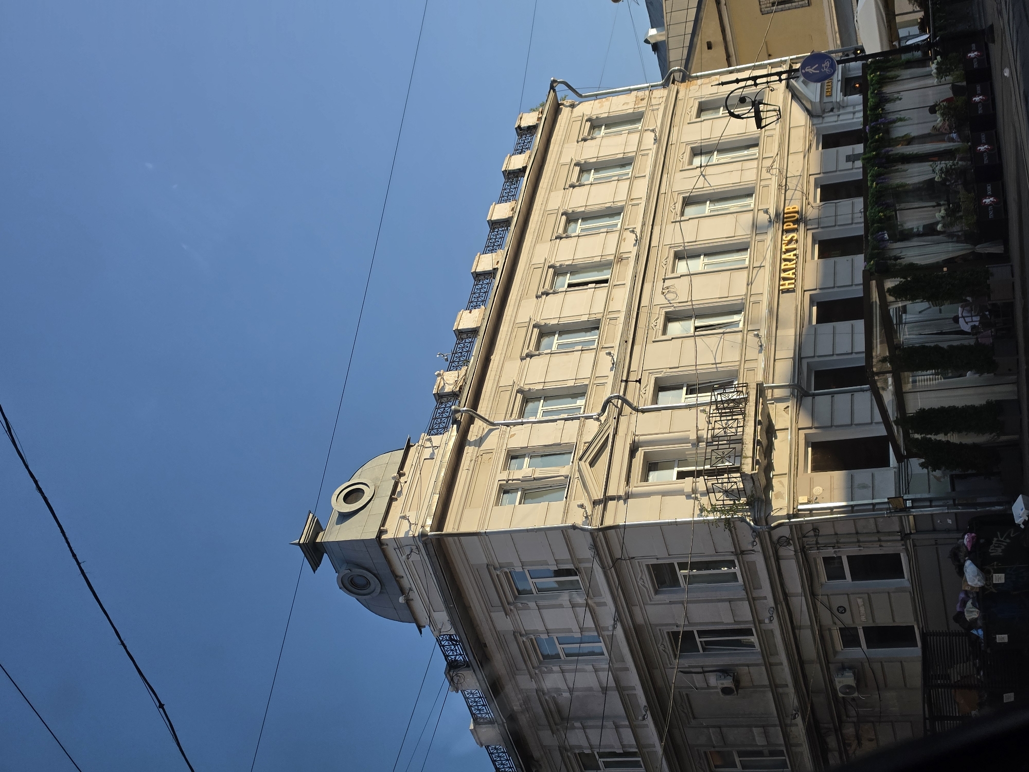 The image captures a street-level view of a building in Nizhny Novgorod, Russia, on a clear, sunny day. The building is a multi-story structure with a light-colored facade, featuring numerous windows. A prominent sign reading "HARAT'S PUB" is visible on the lower section of the building, suggesting a commercial establishment. The building also has architectural details such as a decorative railing along the rooftop and a domed structure on the corner. In the foreground, an outdoor seating area for the pub is set up with tables and greenery. Power lines are visible stretching across the top of the frame against the blue sky. Trash bins are also present at the base of the building. A street sign with a pedestrian and bicycle symbol is mounted on a pole near the pub entrance.