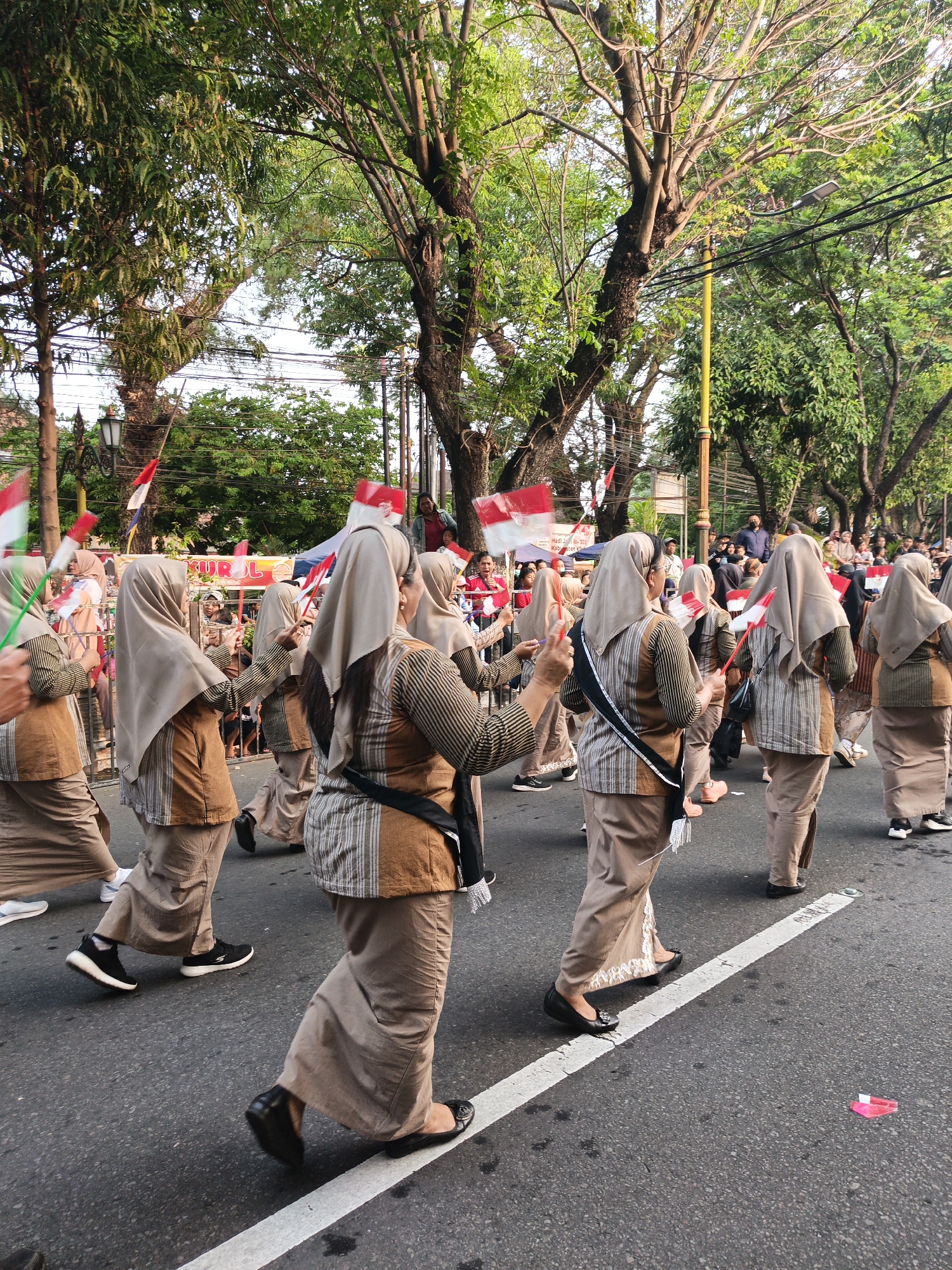 The image depicts a street parade in Klaten, Indonesia, with a group of women walking in unison. They are wearing matching tan and brown traditional clothing, including long skirts and tunics, with light brown headscarves. Several women are holding small red and white flags, which appear to be the Indonesian flag, and some are raising them in the air. The parade is taking place on an asphalt road, with trees and overhead power lines lining the street. Spectators can be seen in the background, some standing behind a barrier. The lighting suggests it is daytime, possibly late afternoon, given the warm tones of the light.
