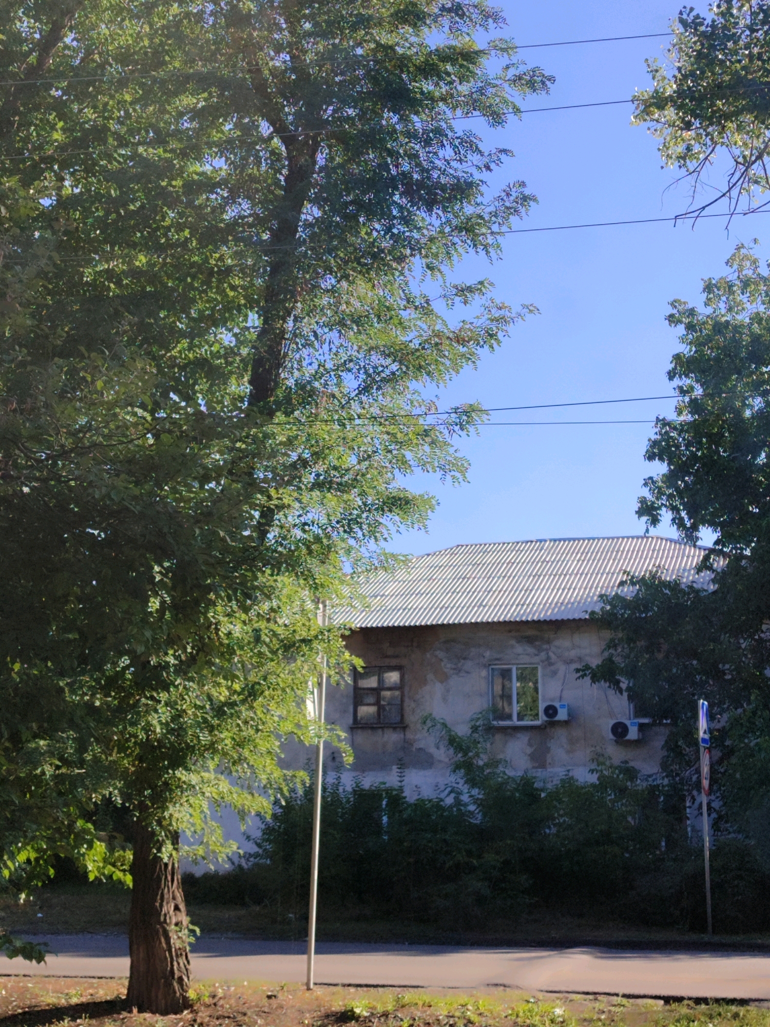The image shows a building with a corrugated metal roof and rough-textured walls, possibly concrete or stucco, in an urban setting. The building has several windows, two of which are visible in the center of the frame. One window is smaller and appears to have wooden frames, while the other is larger and has modern white frames. Two outdoor air conditioning units are mounted on the wall below the larger window. A dense line of green trees and foliage partially obscures the lower portion of the building. In the foreground, a paved road runs horizontally across the bottom of the image, with a grassy area and a mature tree to the left. The sky is clear and blue. A utility pole with power lines runs across the upper part of the image, and a street sign is visible on the right side of the frame.