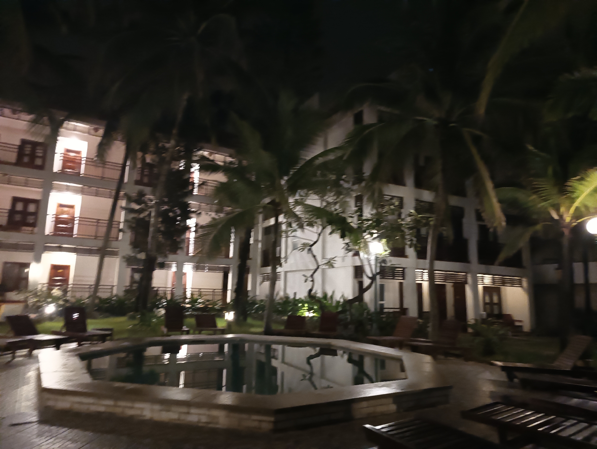 The image depicts a nighttime scene at what appears to be a resort or hotel in Mui Ne, Vietnam. In the foreground, a hexagonal swimming pool is visible, with its water reflecting the lights from the surrounding buildings and palm trees. The pool is bordered by a light-colored coping. Surrounding the pool, there are several lounge chairs, some of which are empty, and some are occupied by indistinct dark shapes that might be shadows or possibly people.
Behind the pool and lounge chairs, a multi-story building complex is visible. The building has a white facade and features multiple balconies with railings and dark-colored doors or windows, many of which are illuminated from within. Tall palm trees are interspersed between the building and the pool area, their fronds creating a dark silhouette against the night sky. The lighting in the scene is a mix of artificial lights from the building and what appear to be outdoor lamps, casting a warm glow on parts of the scene. The overall impression is a tranquil, dimly lit resort atmosphere.