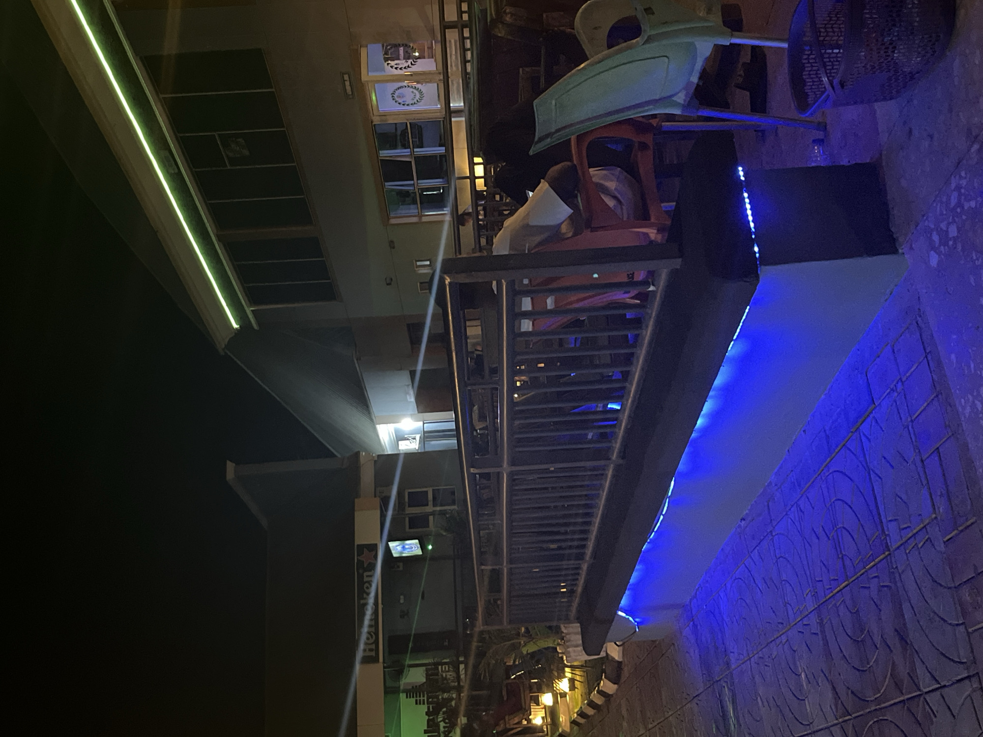 The image shows a nighttime scene in an outdoor seating area of a establishment. A railing runs along the edge of the seating area, illuminated by blue LED lights. Beyond the railing, several chairs are visible, including a red plastic chair and a green plastic chair. In the background, a building with windows and a green neon light strip is illuminated. A person is seated in the red chair, facing away from the camera. The ground is paved with patterned tiles. A Heineken sign is visible on the building to the left. A trash bin is present to the right of the scene.