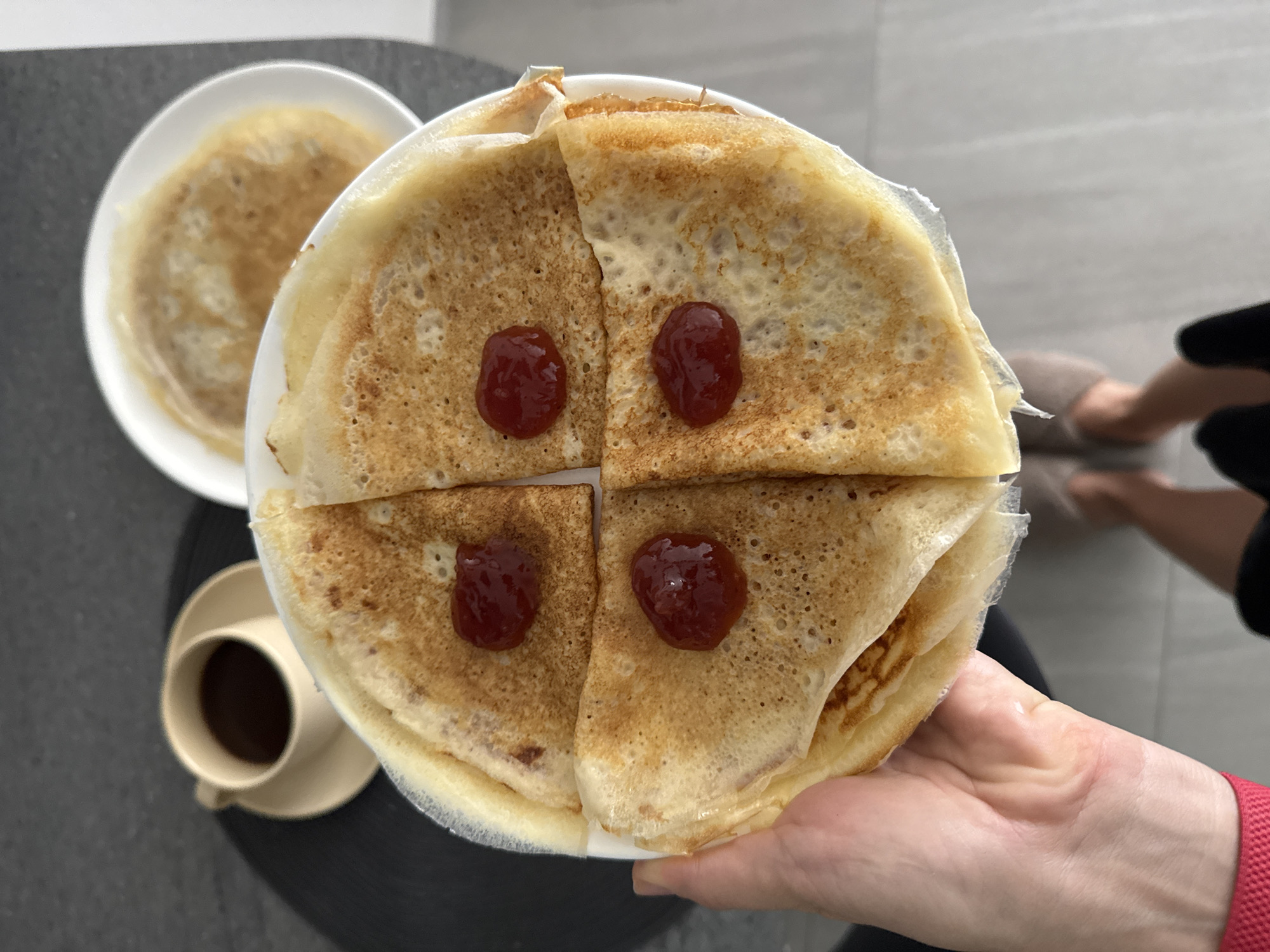 The image shows a first-person perspective of a person holding a plate of folded pancakes topped with red jam. There are four pancakes on the plate, each folded into quarters and adorned with a dollop of jam in the center. A cup of dark liquid (likely coffee or tea) sits on a saucer to the left of the pancake plate, and another plate with a single pancake is behind. A portion of a person's legs and feet, wearing slippers, is visible in the background. The surface under the food and drink appears to be a dark gray or black round table or countertop. Based on the prompt, this scene is supposedly located in Choeng Thale, Thailand.