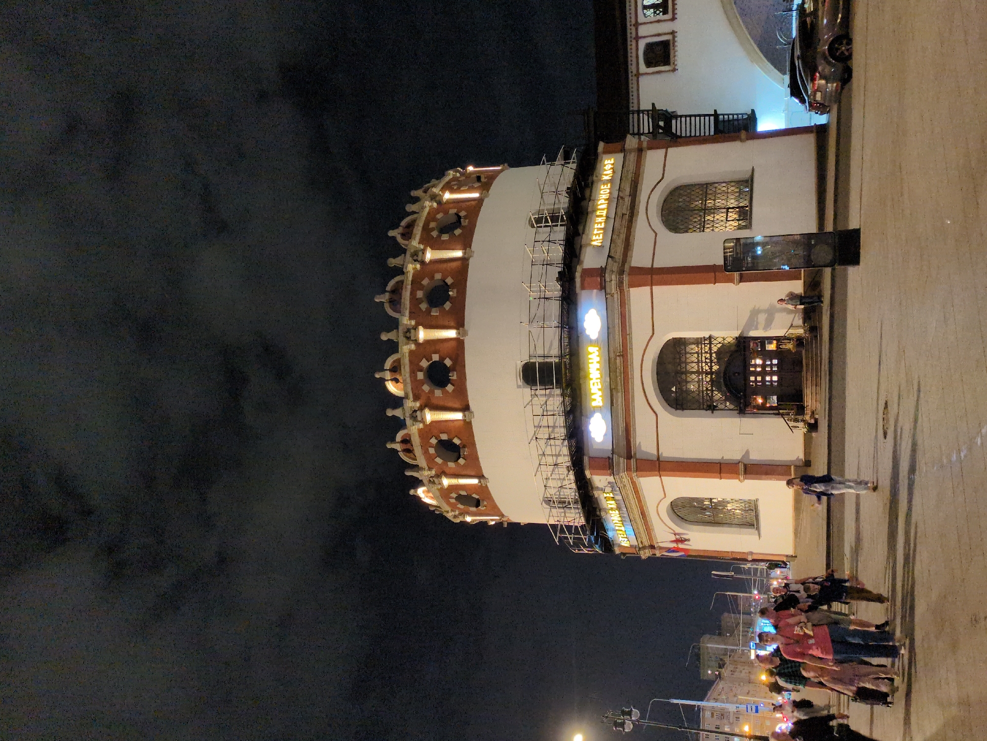 The image depicts a nighttime scene in Moscow, Russia, featuring a distinctive circular building with red and white accents. The building has a decorative top with illuminated circular windows and a white railing around its upper level. On the lower portion of the building, there are large arched windows with intricate metalwork. Yellow neon signs are visible on the building, with one clearly reading "ЛЕГЕНДАРНОЕ КАФЕ" (Legendary Cafe). Several people are walking in the foreground on a paved area, some pulling luggage. A few cars are parked in the background, and streetlights and buildings of the city are visible. The sky is dark and cloudy, characteristic of an urban night.