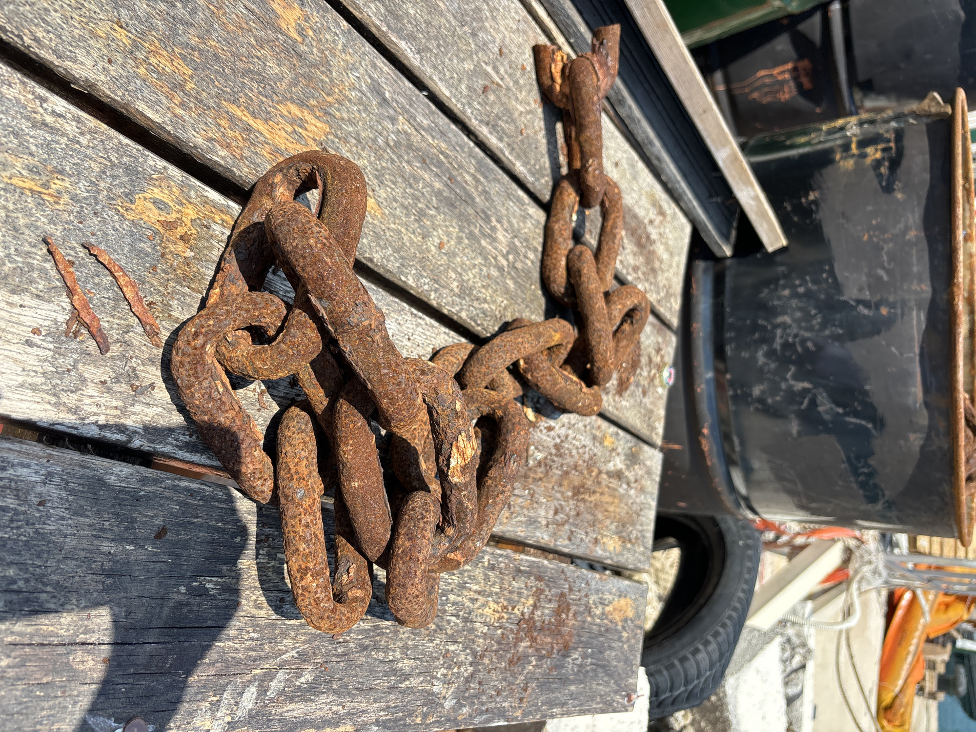 This media file depicts a close-up, overhead view of a rusty metal chain lying on weathered wooden planks. The planks appear to be part of a dock or outdoor platform, suggested by their weathered texture and arrangement. The chain is the primary subject, showing significant signs of rust and age, with some links appearing worn and pitted.
To the right of the chain, a portion of a dark, likely black, object is visible, possibly part of a vehicle or boat, with some reflective surfaces and hints of orange and green colors. A black tire is also partially visible. In the background, there are blurry elements that suggest an outdoor environment with some industrial or marine equipment.
The lighting suggests it is daytime, with strong shadows cast by the chain onto the wooden planks, indicating the sun is relatively high. The scene evokes a sense of disuse or a maritime setting where such chains would be common. There is no visible text in the image. The location context provided is Budva, Montenegro, which aligns with the potential maritime or dockside setting. No people are visible in the media file.