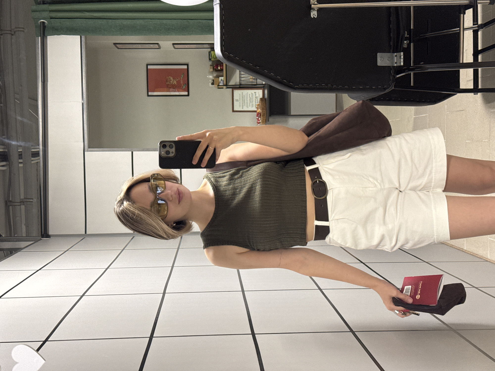 A woman stands in a room with white tiled walls and is taking a mirror selfie. She has blonde hair and is wearing yellow-tinted sunglasses, a dark green ribbed tank top, and white shorts with a dark brown belt. She is holding a passport and keys in her left hand and a brown shoulder bag is slung over her right shoulder. In the background, there is a black chair and a shelf with items. The overall environment appears to be a dressing room or a fitting area.