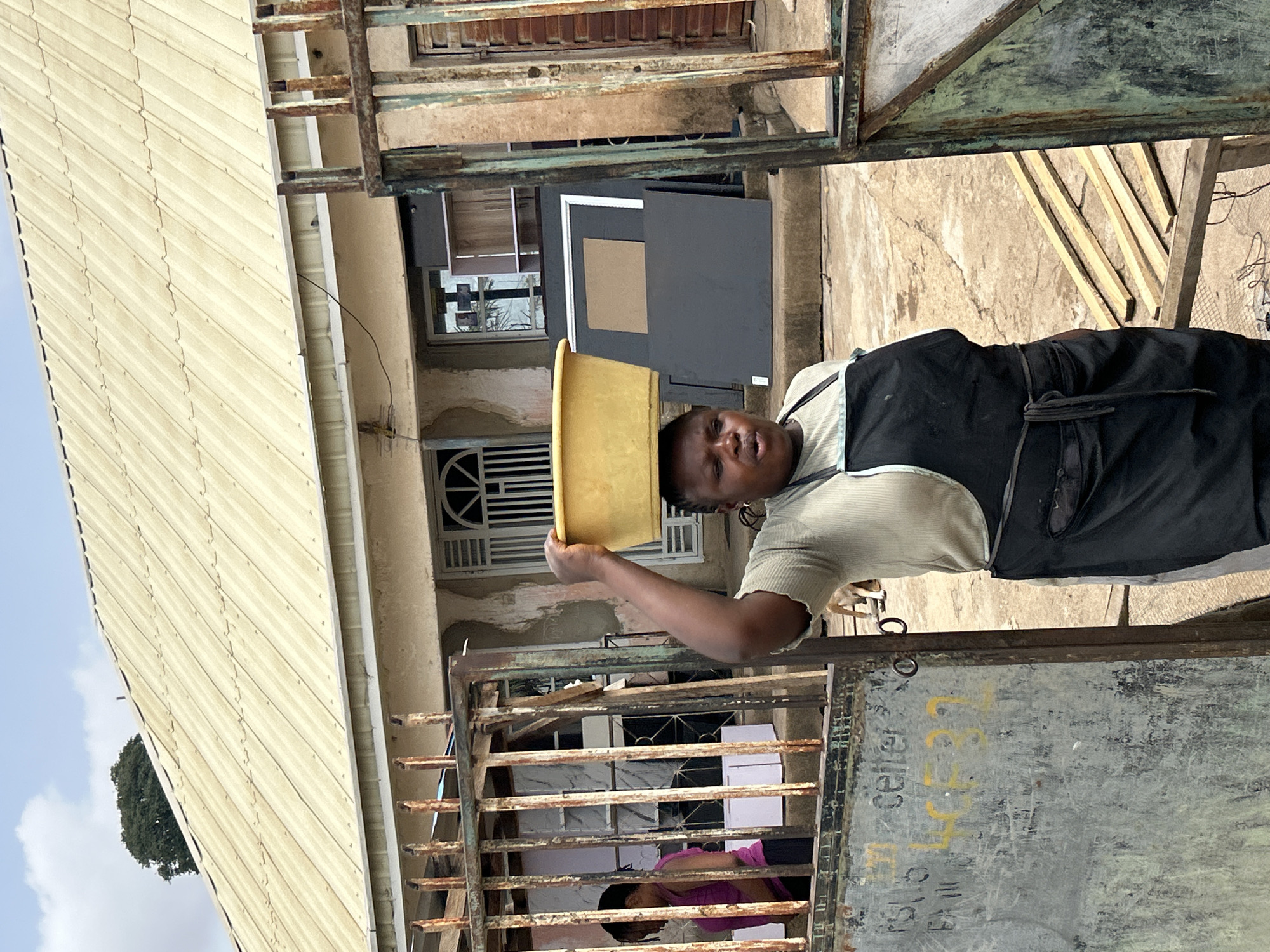 A woman with a yellow bucket balanced on her head stands in front of a building, partly obscured by metal gates with peeling paint and inscribed text. The building has a light-colored, corrugated metal roof and a beige exterior. There's a window with decorative bars and what appears to be an open door or large frame. Another person wearing a pink shirt is visible through the gate. The scene is in Jos, Nigeria.