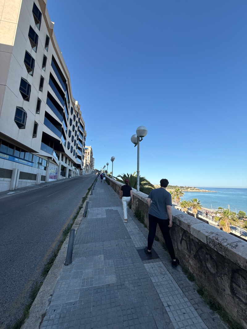A street scene in Tarragona, Spain, shows a downward sloping road and an adjacent elevated pedestrian walkway with a low stone wall. The walkway is paved with square gray tiles and is partially lined with metal bollards along the road edge. Two people are visible on the walkway: one wearing black pants and a gray shirt leaning on the wall facing toward the Mediterranean Sea, and another in white pants and a black shirt walking in the same direction. A large, multi-story building with an elongated, slightly curved facade and distinctive, deep-set windows lines the road, on the left. The Mediterranean Sea is visible in the distance, lined with palm trees and other buildings. The sky is clear and blue. Street lights are spaced along the walkway.