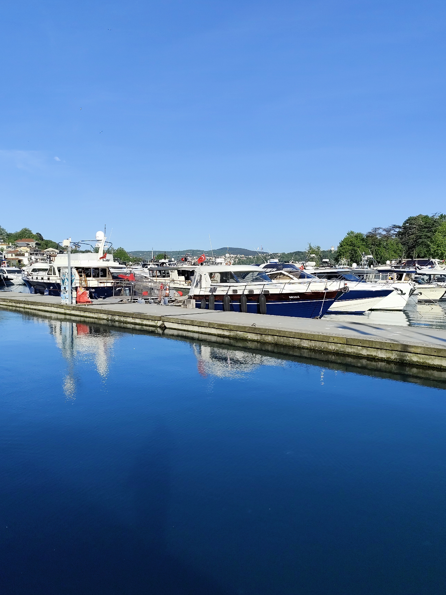 The image depicts a marina with several boats docked along a concrete pier in Sarıyer, Turkey. The water is deep blue and reflects the boats and sky. The boats vary in size and design, with some featuring white hulls and others with darker blue or brown accents. One boat is labeled "MONI". Green trees and residential buildings are visible in the background. The sky is clear and blue.