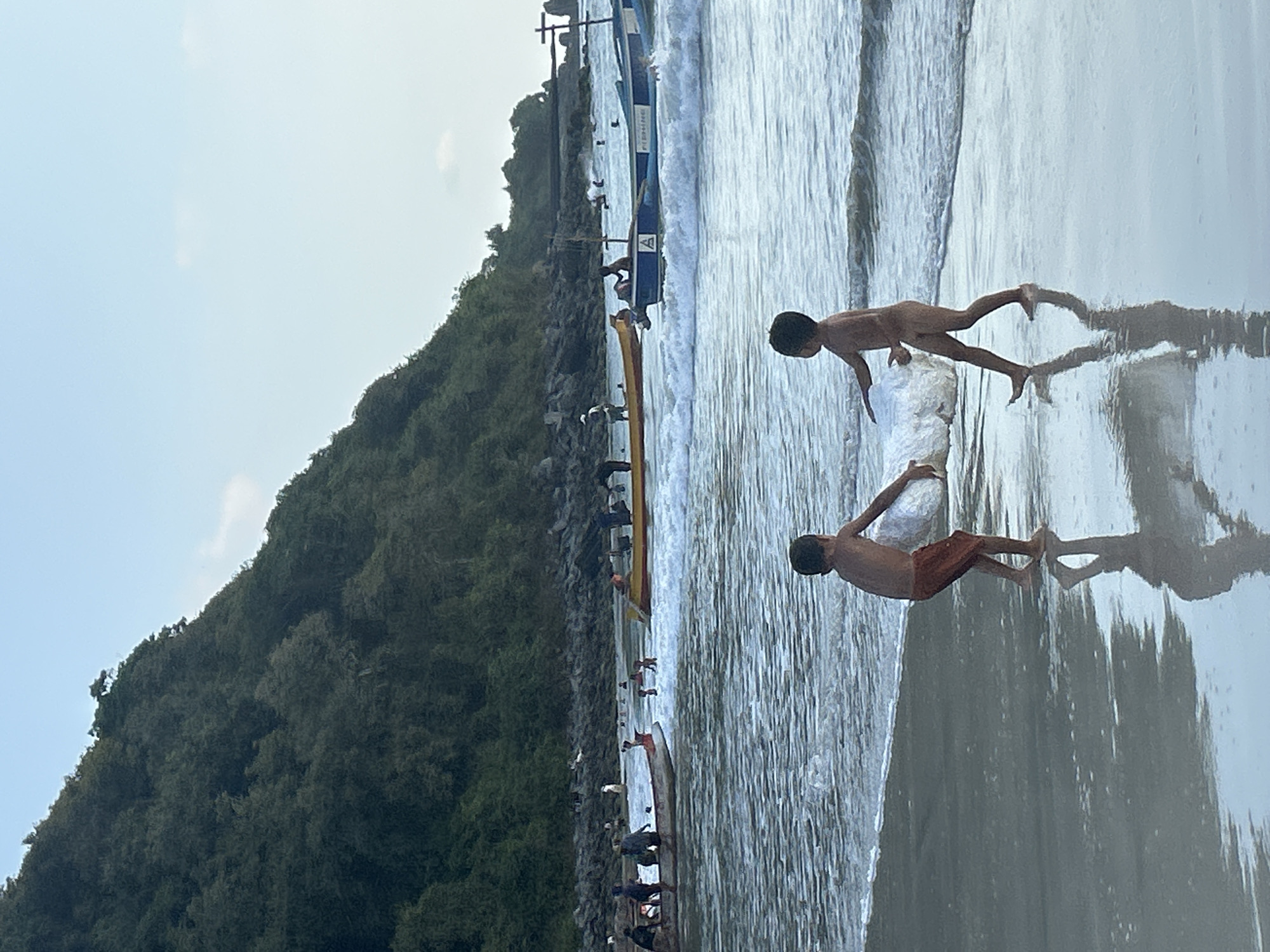 The image depicts a lively scene at Selongbelanak beach in Indonesia. Two young boys are the primary subjects, one in the foreground and the other further back. Both are partially submerged in the shallow ocean water. The boy in the foreground is leaning forward, appearing to interact with a large, white, amorphous object. The second boy is positioned behind him, also looking towards the object. Their bodies are wet, and they are dressed in swimwear.
The setting is a beach with gentle waves lapping the shore. The water is a light blue-gray, reflecting the sky. To the left of the frame, a lush green, tree-covered hillside rises steeply, suggesting a coastal environment with natural topography. Along the waterline, a line of traditional Indonesian outrigger boats is visible, some with people in or around them. The boats are narrow and long, typical of those used for fishing or transport in the region.
The activity appears to be playful, with the boys seemingly engaged in some sort of game or activity with the object in the water. The presence of the boats and people along the shore indicates a community or gathering at the beach.
The sky is overcast, with hints of diffused sunlight, suggesting it is daytime, possibly late morning or afternoon. The overall mood appears relaxed and casual, typical of a beach environment. The water is calm, indicating pleasant weather conditions.
There is some visible text on one of the boats, appearing as blue lettering. One portion of the text is visible as "A" followed by some smaller letters. Another boat has text that seems to read "BINAGRA" and below it "PURA", indicating it is likely a local fishing or transport boat.
The image captures a slice of everyday life at a beautiful Indonesian beach, highlighting the interaction of people with their environment and local traditions.