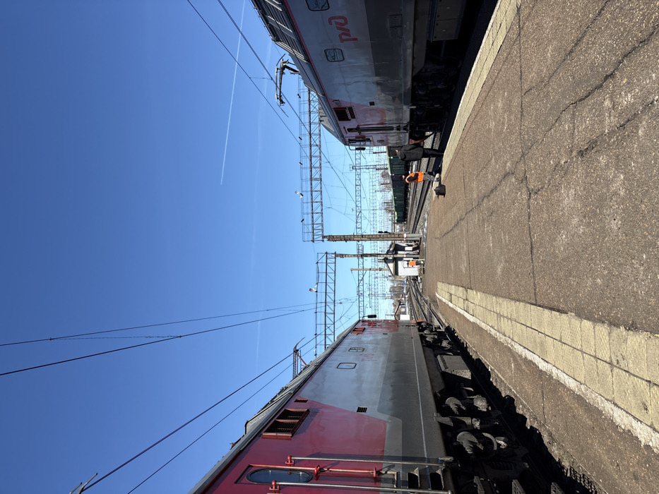 The image depicts a railway station or yard in Kropachyovo, Russia, under a clear, bright blue sky with a single faint contrail. Two large electric locomotives frame the scene, parked on parallel tracks alongside a concrete platform. The locomotive on the left is predominantly red and grey, while the one on the right is grey and red, displaying the "РЖД" (RZD) logo.
The platform, made of cracked concrete with faded yellow tactile paving along its edge, extends into the foreground. Multiple railway tracks converge towards the horizon in the middle ground, featuring extensive overhead electrification lines supported by metal gantries and poles. A small, light-colored utility or signal building stands between the tracks in the mid-distance. Further beyond, green freight cars are visible on additional tracks.
Three individuals are present on the platform. On the right, one person in an orange high-visibility vest stands near the grey and red locomotive, facing towards the tracks. A dark bag rests on the platform nearby. Adjacent to this individual, another person in dark attire stands with their back to the viewer, also near the locomotive. A third person, also in an orange high-visibility vest, is positioned near the small building in the middle distance, facing generally towards the right side of the frame.