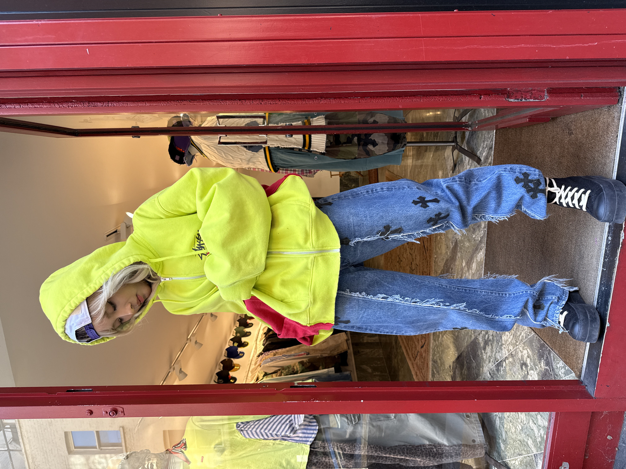 A person stands upright within a prominent red-framed doorway, likely the entrance to a retail store in London, United Kingdom. The individual has light-colored hair and is dressed in a vibrant, casual outfit. They wear a bright neon yellow zip-up hoodie, featuring the "STUSSY" logo on the left chest, layered over a red garment. A white baseball cap, also bearing the "STUSSY" logo, is visible beneath the hoodie's hood, and sunglasses are pushed up onto their head. They are wearing blue, heavily distressed jeans with numerous black cross-shaped patches or appliques, paired with chunky black high-top shoes secured with white laces.
The person is depicted looking downwards with a calm expression, their arms tucked into the hoodie. The red door frame, showing some wear, provides a bold contrast to the person's attire. Through the glass of the door, the interior of a clothing store is visible, displaying racks of clothing including a camouflage jacket and a striped shirt, as well as shelves stocked with hats. A reflection of an exterior building with a window indicates it is daytime. The flooring within the doorway appears to be a combination of patterned tiles and a darker mat.