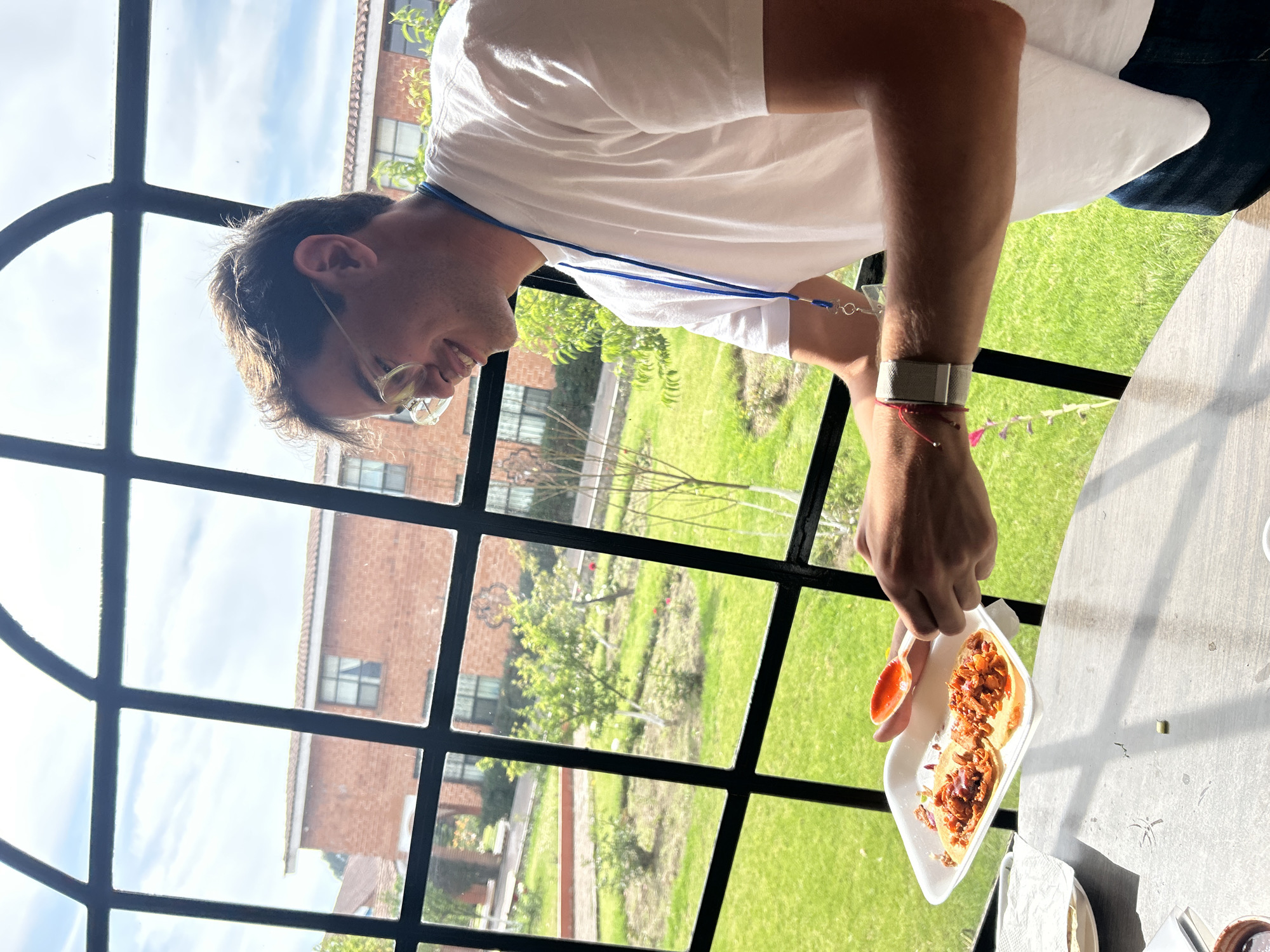 A man with short brown hair and glasses is seen in a medium shot, wearing a white t-shirt and a blue lanyard. He is looking down and slightly to the left, smiling. He is holding a white Styrofoam container with what appears to be two tacos and a small dish of red sauce. He is about to add the sauce to the tacos. The background shows a large, arched window with black grilles, through which a view of a green lawn, trees, and a brick building can be seen. The setting is indoors, with a table partially visible in the foreground. The city and state are Unknown, Unknown.