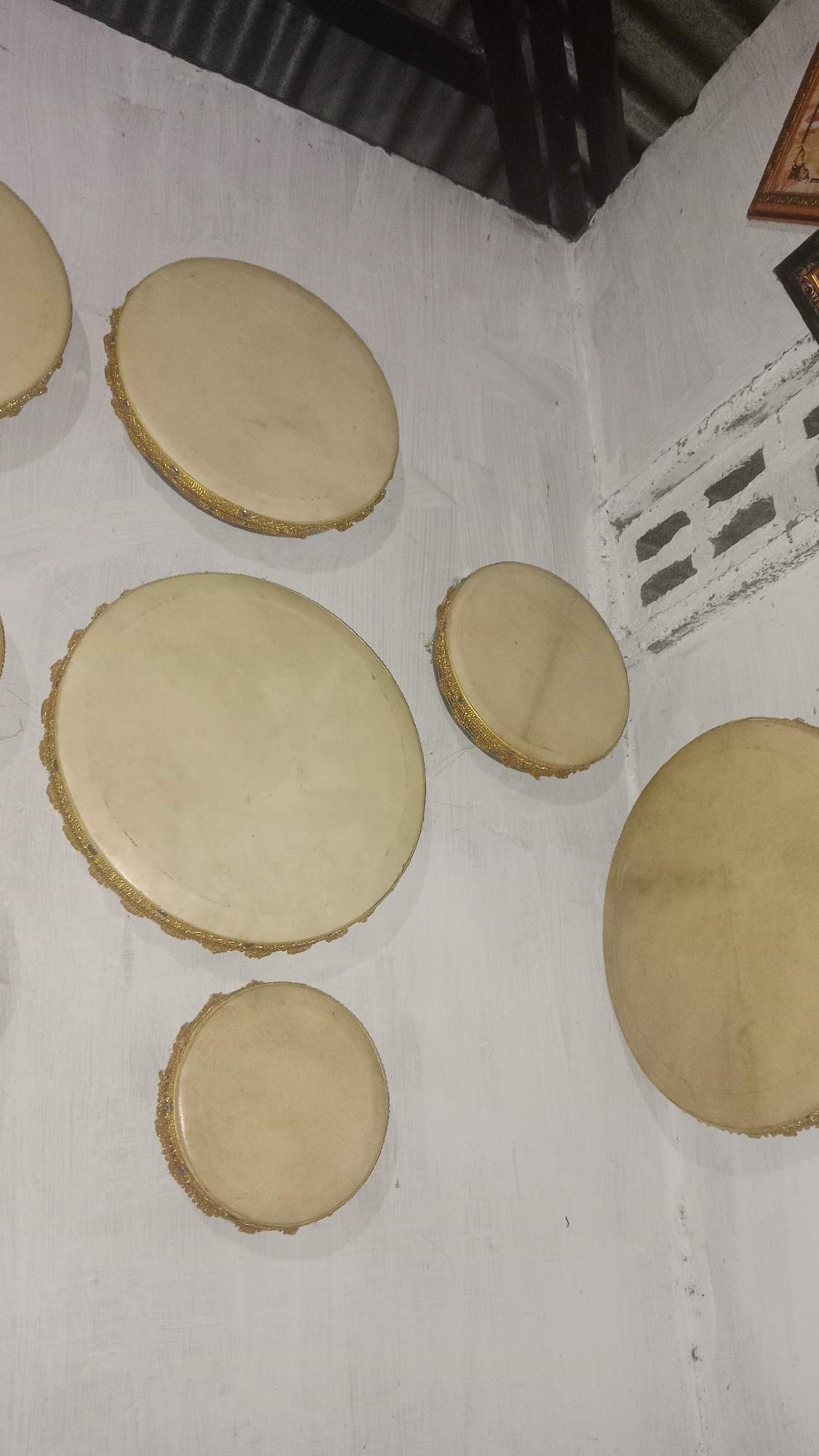 The image shows several tan-colored frame drums, also known as tambourines or duffs, mounted on a white, textured wall. The drums vary in size and are arranged in a staggered pattern. They all appear to have a tan or light brown membrane stretched across their circular frames, which are decorated with gold trim. The wall itself has visible brush strokes and appears to be indoors, possibly in a room with a corrugated metal ceiling visible in the upper portion of the image. A glimpse of what appears to be a framed picture can be seen in the upper right corner. The scene suggests a decorative display of musical instruments in a simple setting.