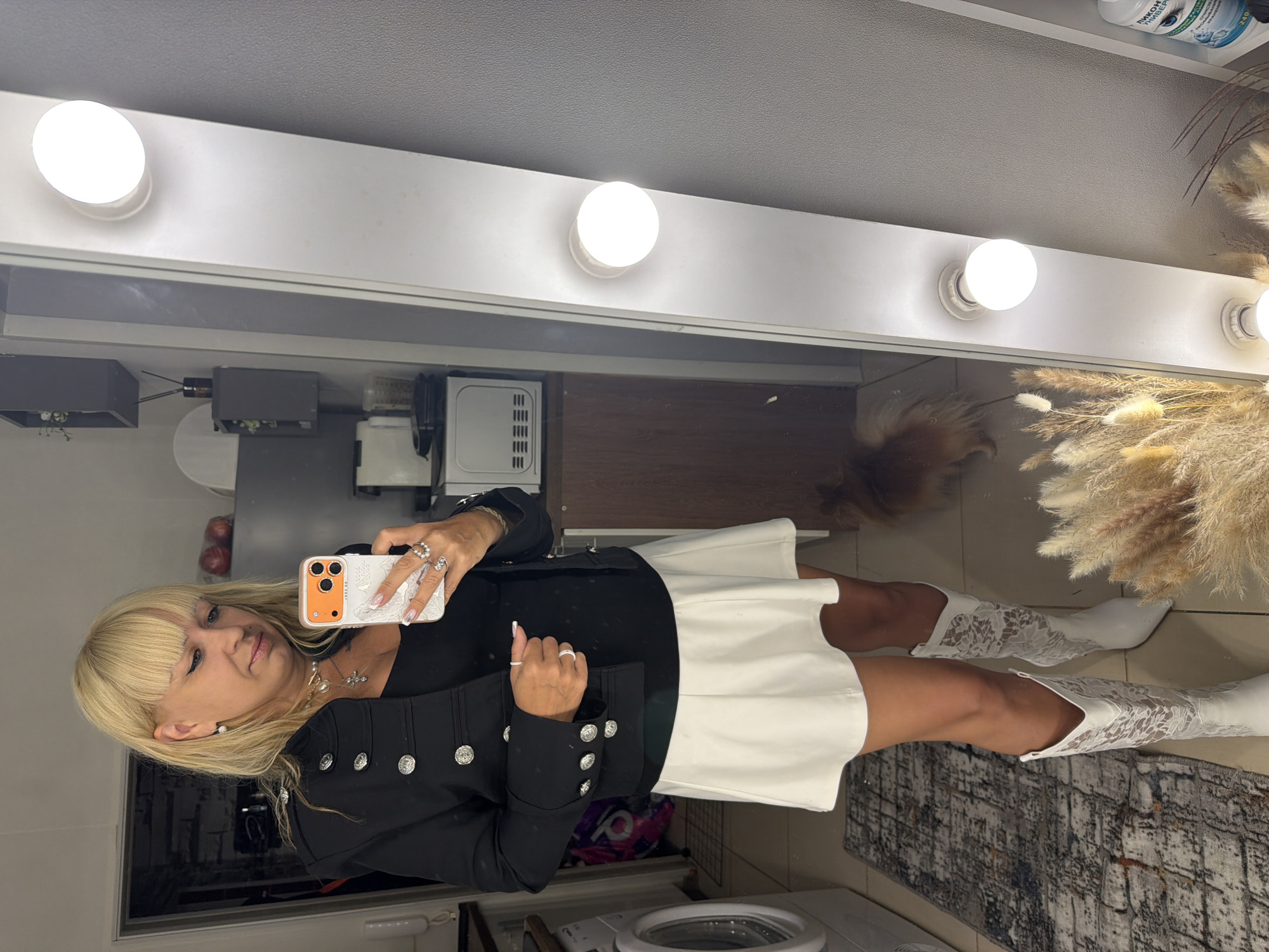 A woman is taking a selfie in a bathroom or dressing room, characterized by bright vanity lights above a mirror. She has blonde hair and is wearing a black jacket with silver buttons, a white skirt, and white lace boots. She is holding a white and orange smartphone in her hand. The background includes a washing machine, countertops with various items like a microwave, flowers, and decorative pampas grass. The location is indicated as Chaikovsky, Russia, likely within a residential setting.