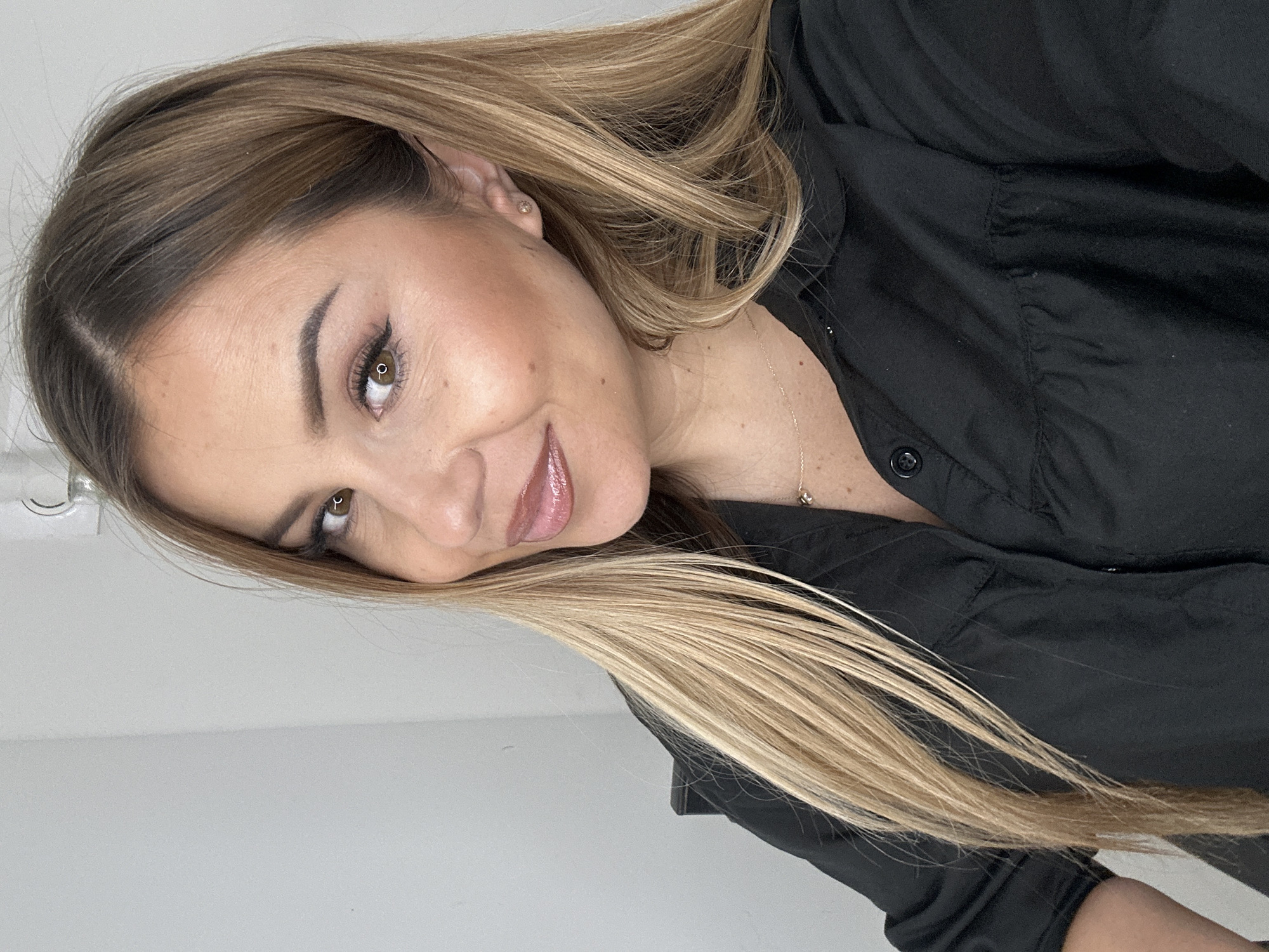 A woman with long blonde and brown ombre hair is shown in a medium close-up, looking towards the viewer. She is wearing a black collared shirt. Her makeup is visible, including eyeliner and lip gloss. She has a small stud earring in her visible ear and a delicate necklace. The background is a plain, light-colored wall. The lighting is soft and even, suggesting an indoor setting. There is no indication of the location in Asenovgrad, Bulgaria.