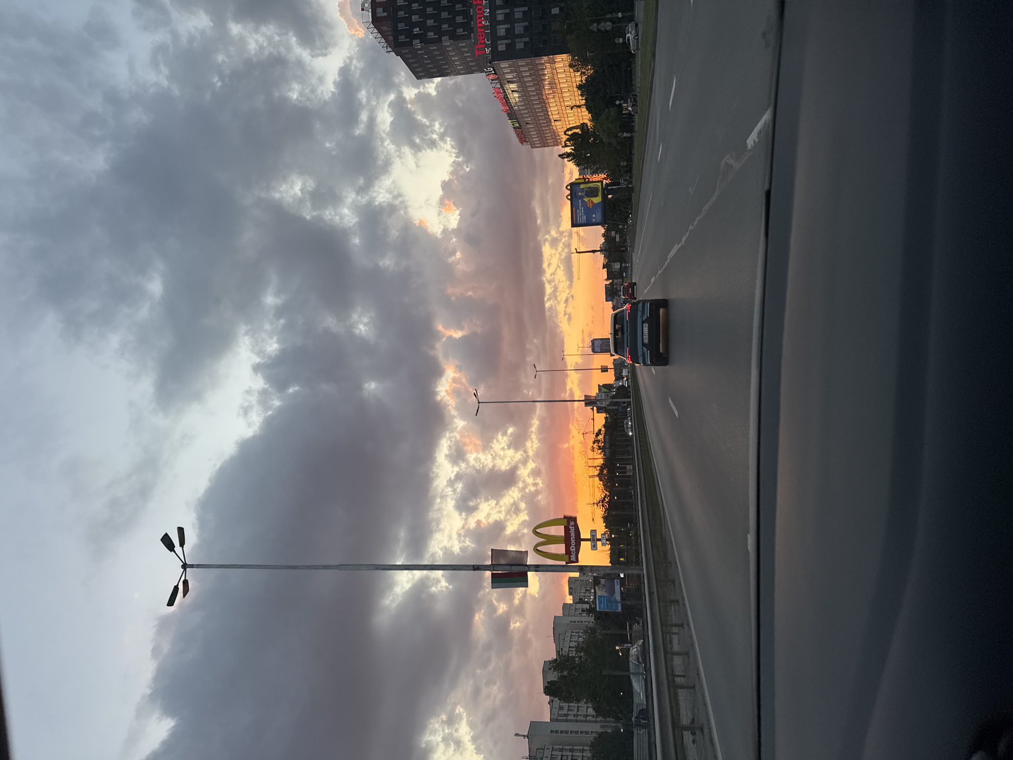 The image presents a forward-facing view from inside a vehicle on a multi-lane urban highway during sunset in Sofia, Bulgaria. The sky is largely overcast with dark grey clouds, but the horizon is illuminated by a vibrant band of orange, pink, and yellow light from the setting sun, particularly pronounced on the right side of the frame.
In the immediate foreground, the dark grey dashboard and the lower edge of the windshield of the photographing vehicle are visible.
The central subject is a paved highway with at least four lanes of traffic moving away from the camera. White lane markings are discernible, with some areas showing wear. A metal guardrail runs along the left side of the road, separating the main carriageway from a possible service road or a shoulder.
Ahead in the central lane, a dark blue or teal Audi e-tron SUV is prominently featured, displaying a Bulgarian license plate "CB 8311 OM". Further ahead, a red sedan is also visible in the same lane. Other vehicles are present in the distant lanes, mostly obscured by distance and lighting.
On the left side of the highway, a tall grey lamppost with four multi-directional light fixtures stands prominently. Below the fixtures, a banner displaying the vertical green, white, and red stripes of the Bulgarian national flag is affixed. Further down the road on the left, a large yellow McDonald's "M" logo sign is clearly visible. Behind these features, several multi-story, light-colored residential apartment blocks with numerous windows line the street. A blue billboard is also visible on this side.
On the right side of the highway, lush dark green trees and bushes border the road. Beyond the foliage, a large, modern, dark-clad high-rise building with extensive glass facades is prominent. Its windows brightly reflect the orange and yellow light of the sunset. White text signs for "Thermo Fisher Scientific" and "LG" are visible on the building's exterior.
Based on the Bulgarian flag, the "CB" prefix on the license plate (designating Sofia City), the distinctive McDonald's branding, the architecture (both the older residential blocks and the specific modern high-rise bearing "Thermo Fisher Scientific" and "LG"), the location is identifiable as **Tsarigradsko Shose Boulevard in Sofia, Bulgaria**, likely looking towards the southeast, with the modern building being Capital Fort.