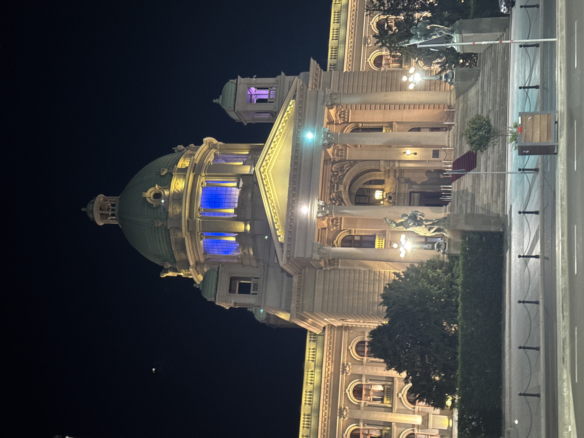 The image shows the National Assembly of Serbia in Belgrade, Serbia, at night. The building is lit with warm lights, and the dome is illuminated with blue and purple lights. The street in front of the building is visible, as are trees and statues. The sky is dark.