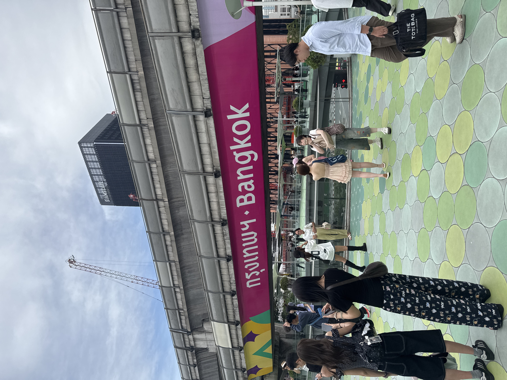 The image depicts an elevated walkway in Bangkok, Thailand. The walkway has a patterned green and yellow circular tile floor. A large magenta sign reads "Bangkok" in English and Thai. People are walking and standing on the walkway. A man in a light blue shirt and brown pants carries a black tote bag with "THE TOTE BAG" printed on it. Several women are visible, wearing dresses, skirts, and pants. A skyscraper with the name "SIAMS" is visible in the background. A construction crane is also visible. The sky is cloudy.