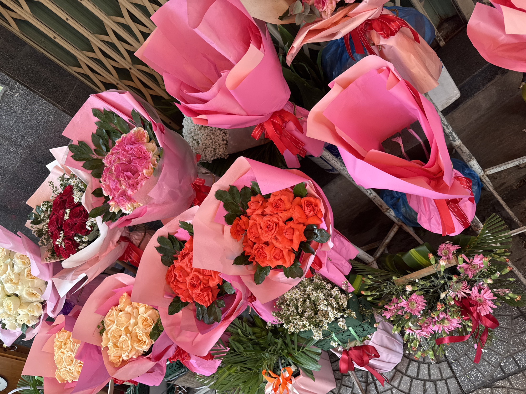 The image captures a collection of flower bouquets laid out on a sidewalk in what appears to be Đà Nẵng, Vietnam. The bouquets, wrapped in pink paper and tied with ribbons, showcase a variety of flowers including roses in shades of red, orange, and peach, as well as mixed bouquets featuring pink and white flowers. Some bouquets also include greenery and baby's breath. The bouquets are displayed on the ground, possibly for sale. The sidewalk is made of stone tiles and in the background there is a metal gate.