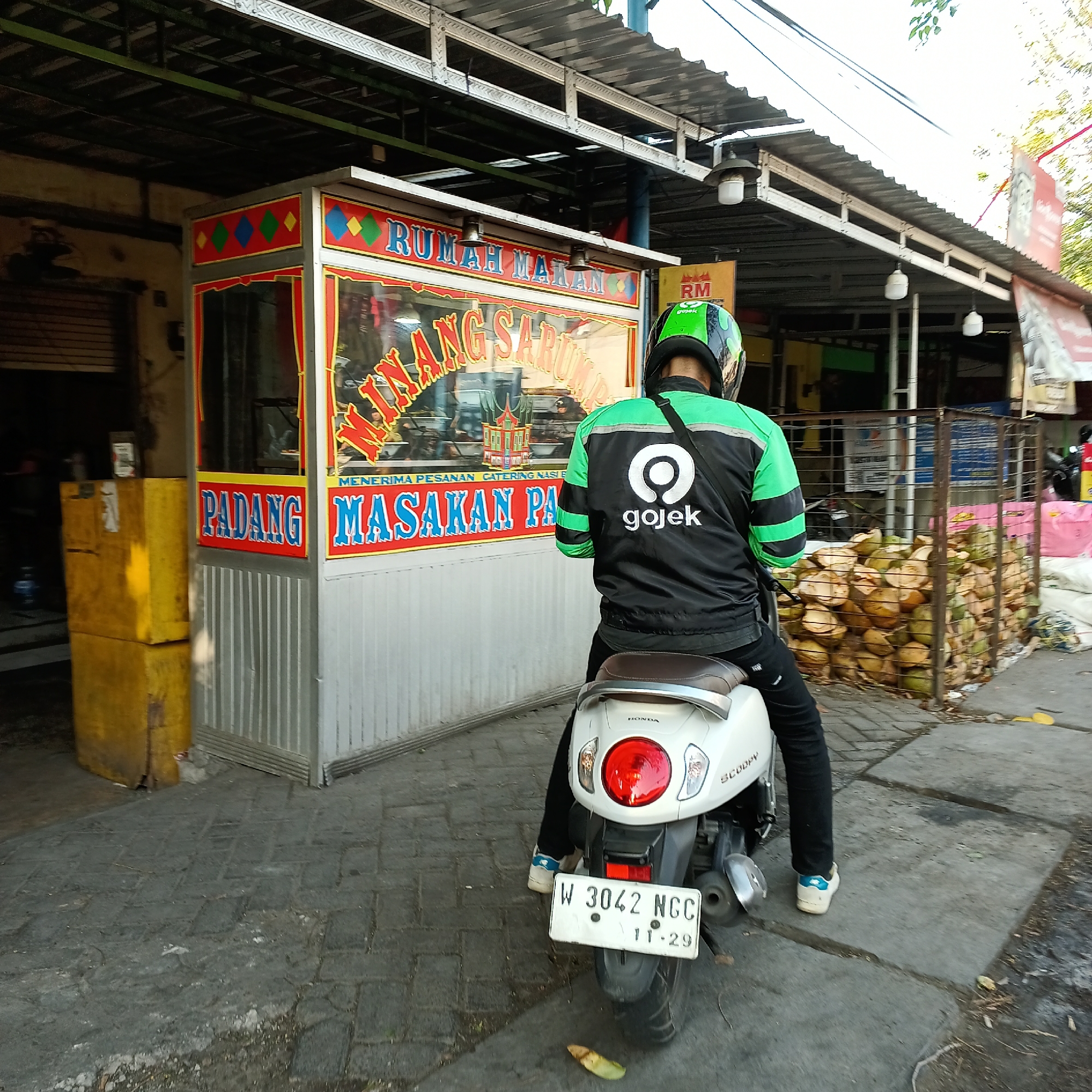 In Wedoro, Indonesia, a Gojek delivery driver, identified by his green and black jacket with the Gojek logo, sits on a white Honda Scoopy motorcycle. The driver is facing away from the camera, looking towards a food stall. The food stall, labeled "RUMAH MAKAN" (meaning "Eating House") and "PADANG MASAKAN PA" (indicating "Padang Cuisine"), has a colorful facade with diamond patterns and text indicating that catering services are accepted. Inside the stall, food is visible behind a glass display. To the right of the food stall, a cage is filled with cut coconuts. The scene is set on a paved walkway with some foliage and shadows suggesting an outdoor setting. The motorcycle's license plate reads "W 3042 NGC 11-29".