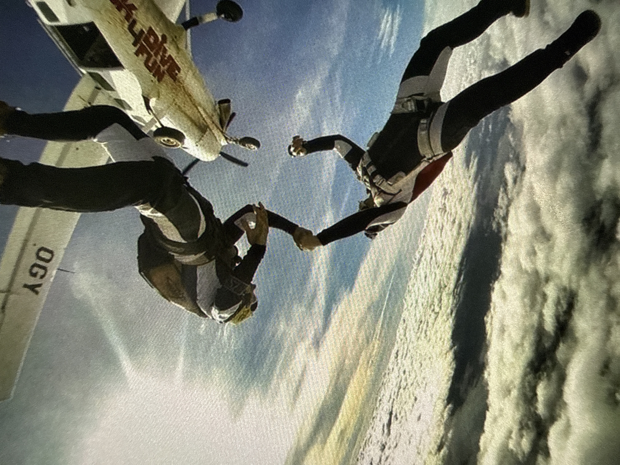 Two skydivers are freefalling while holding hands in front of a small airplane. The airplane, with the words "SKY DIVE FUN" visible on its side, is flying at an angle relative to the divers. A wing with the letters "OGY" is visible on the other side of the plane. The background features a blue sky above a layer of clouds or snow-covered ground. The location is Unknown, Unknown.