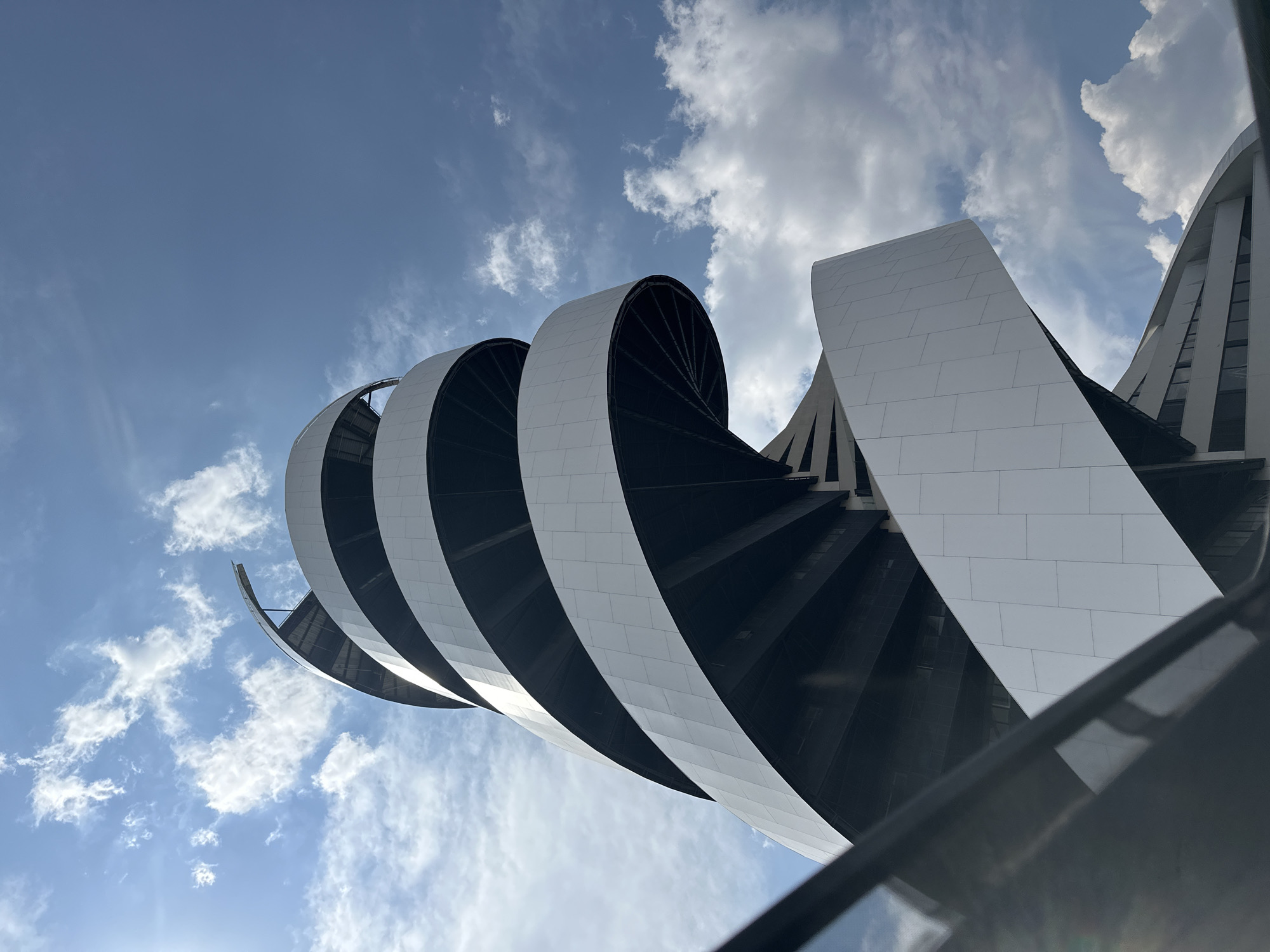 The image depicts a low-angle view of a modern architectural structure with a distinctive spiral design. The building features alternating white and dark sections, creating a striking contrast. The white sections appear to be composed of flat, rectangular panels. The spiral structure is set against a backdrop of a bright blue sky with scattered white clouds. The composition emphasizes the upward-reaching form of the building, with a partially visible dark object in the foreground. The location is San Bernardino Tlaxcalancingo, Mexico.