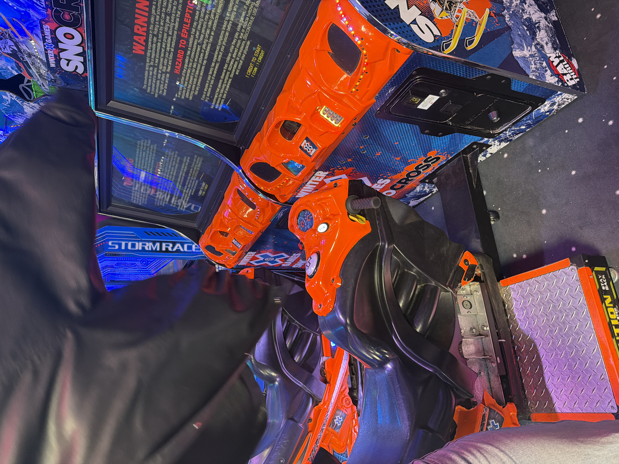 The image shows a "Winter X Games Snocross" arcade game, featuring two snowmobile racing seats with orange and black accents. The game's screens display warnings about photosensitive epilepsy. The floor has a starry pattern. No specific location within Sofia, Bulgaria can be identified.