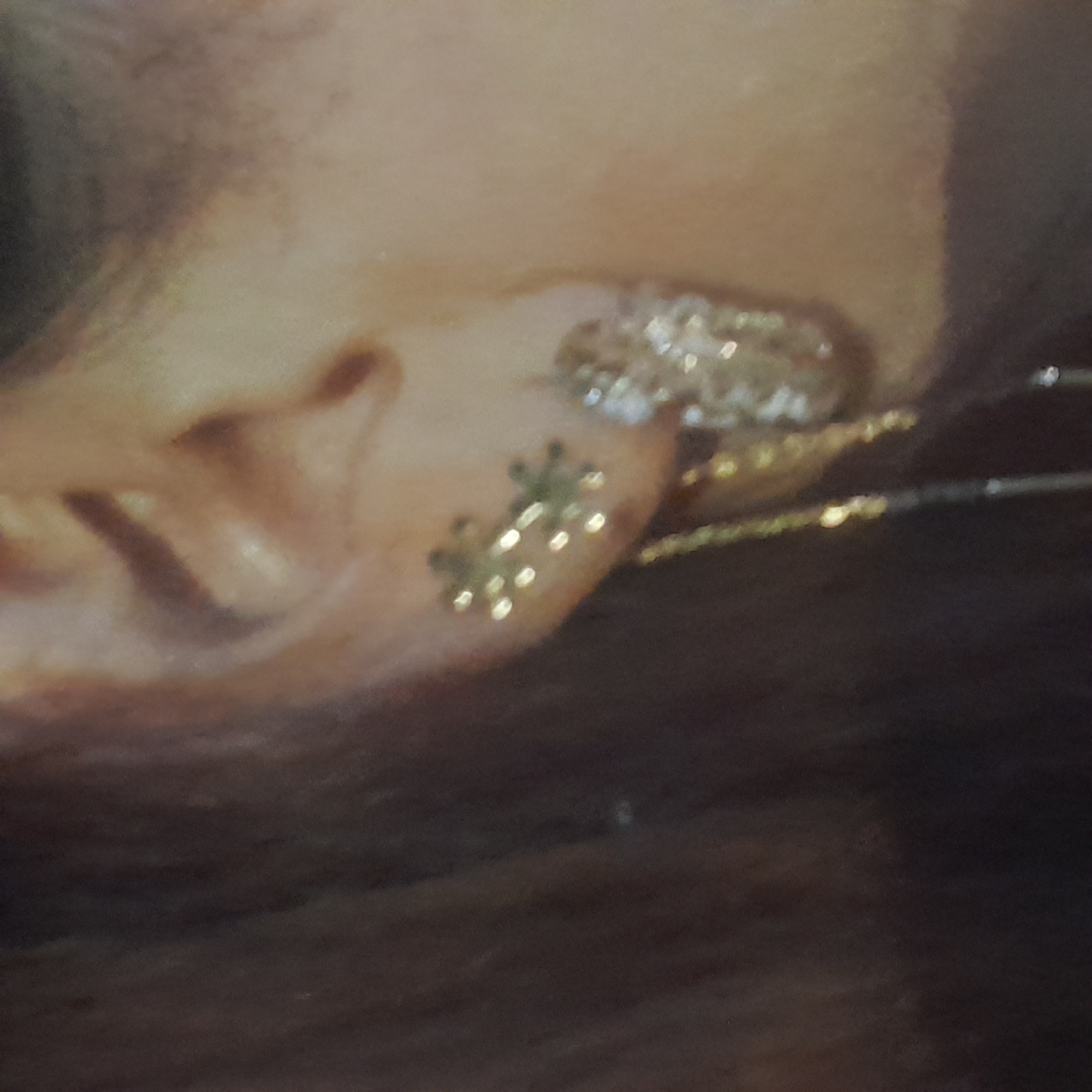 The image shows a close-up of a person's ear with two earrings. The earring on the left is a small, star-shaped stud with multiple points. The earring on the right is larger and appears to be a dangle earring with a cluster of sparkling stones. A thin chain hangs from the right earring. The background is out of focus, with dark hair visible on the left and skin tones on the right. The image does not provide any information about the location in Rudəkənar, Azerbaijan.